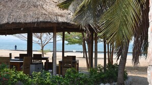 Ein Sitzbereich am Strand mit einem Reetdach, Holzmöbeln und Blick aufs Meer.