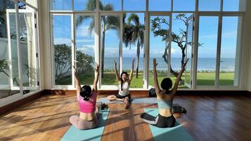 Tres personas practicando yoga sobre colchonetas en una habitación con ventanas amplias que dan a una playa y palmeras.