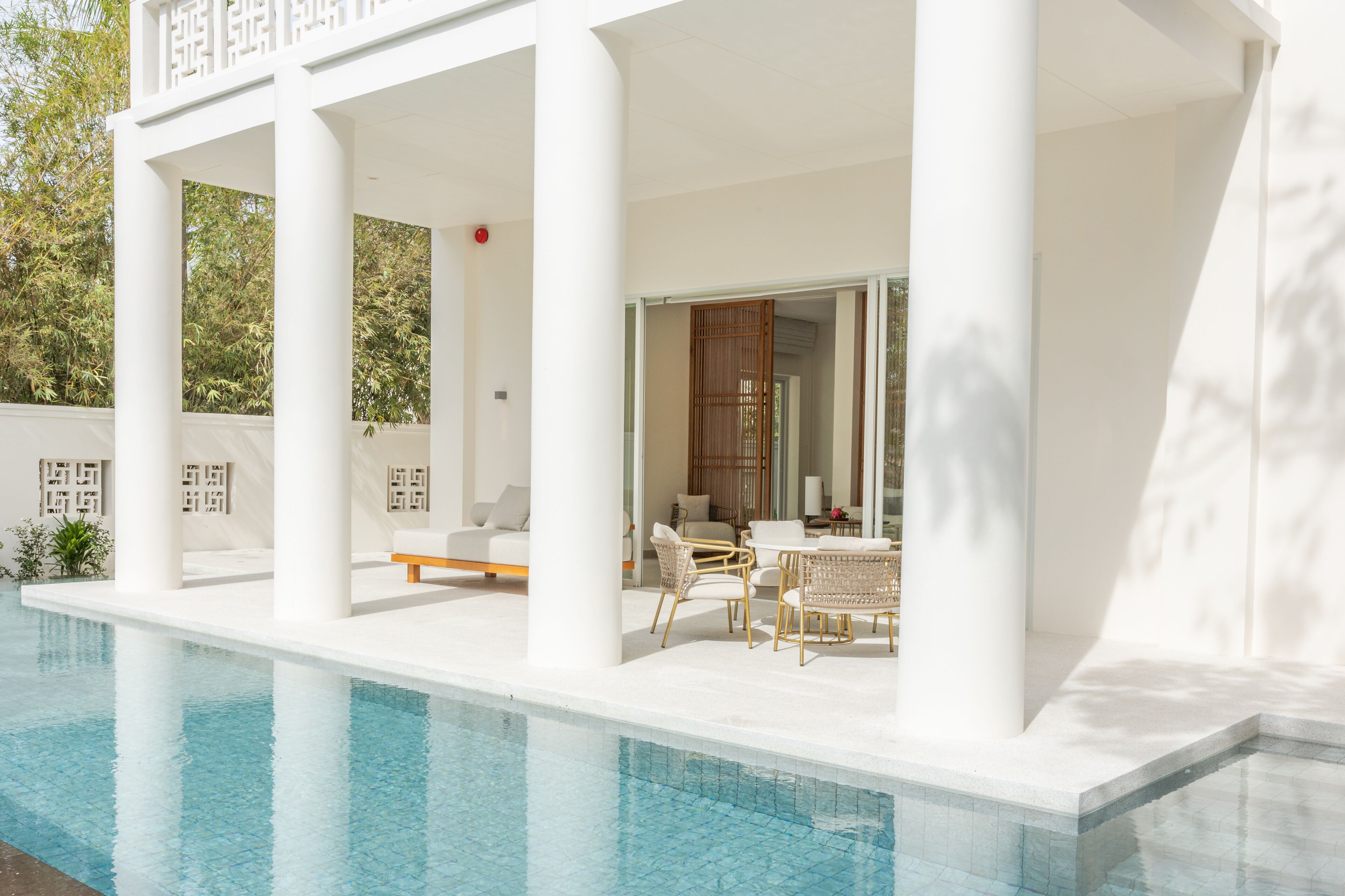 A modern poolside area with white columns, a seating area with wicker furniture, and a view of the pool.