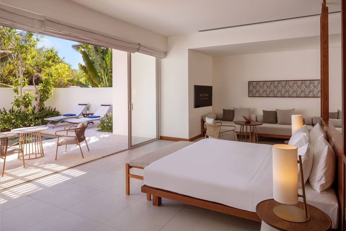 A modern bedroom with a large bed, a sofa, a small table, and a chair. There is a sliding glass door leading to a patio with a pool and lounge chairs.