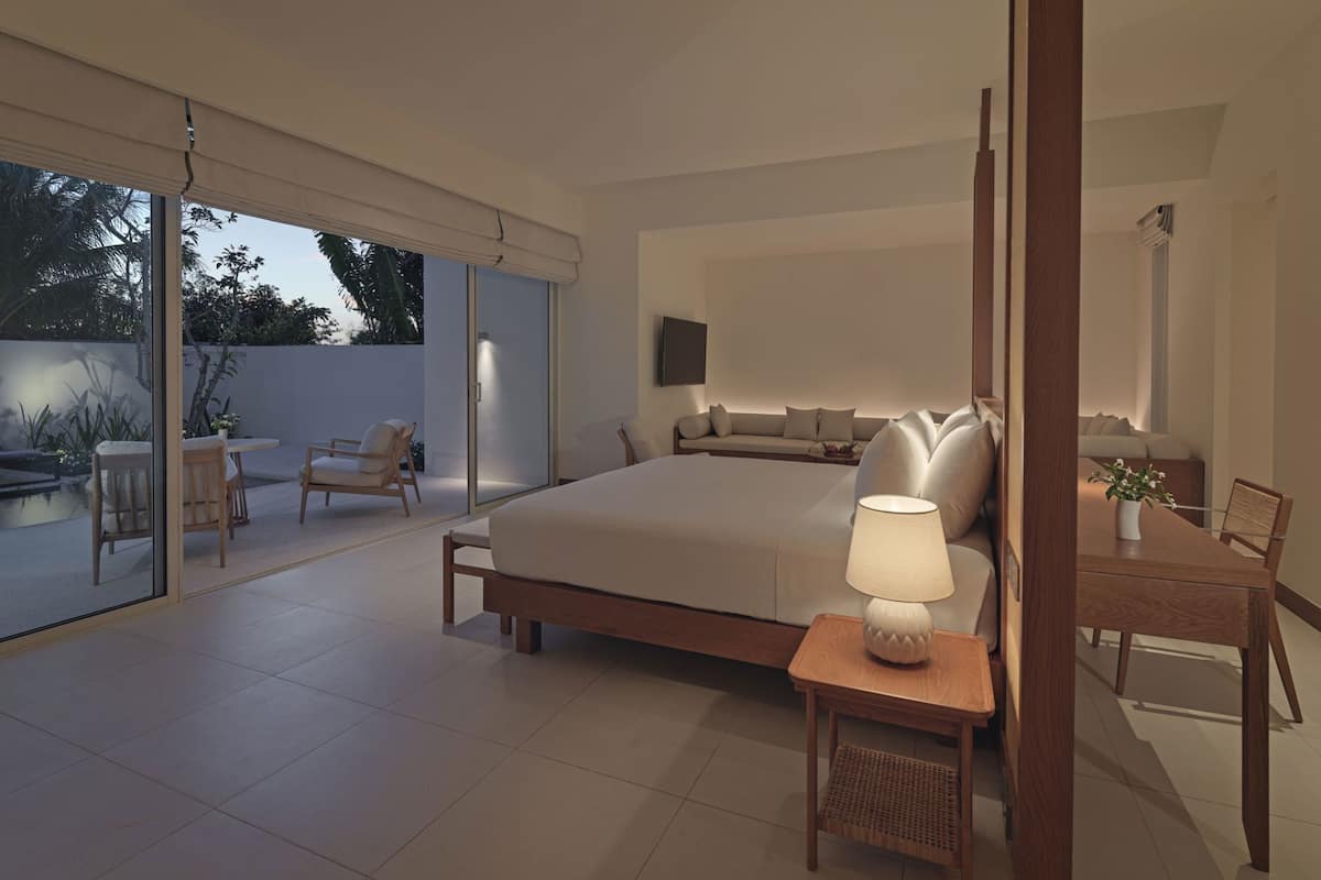 A modern bedroom with a large bed, a bedside table with a lamp, a sofa, and a dining area with chairs.