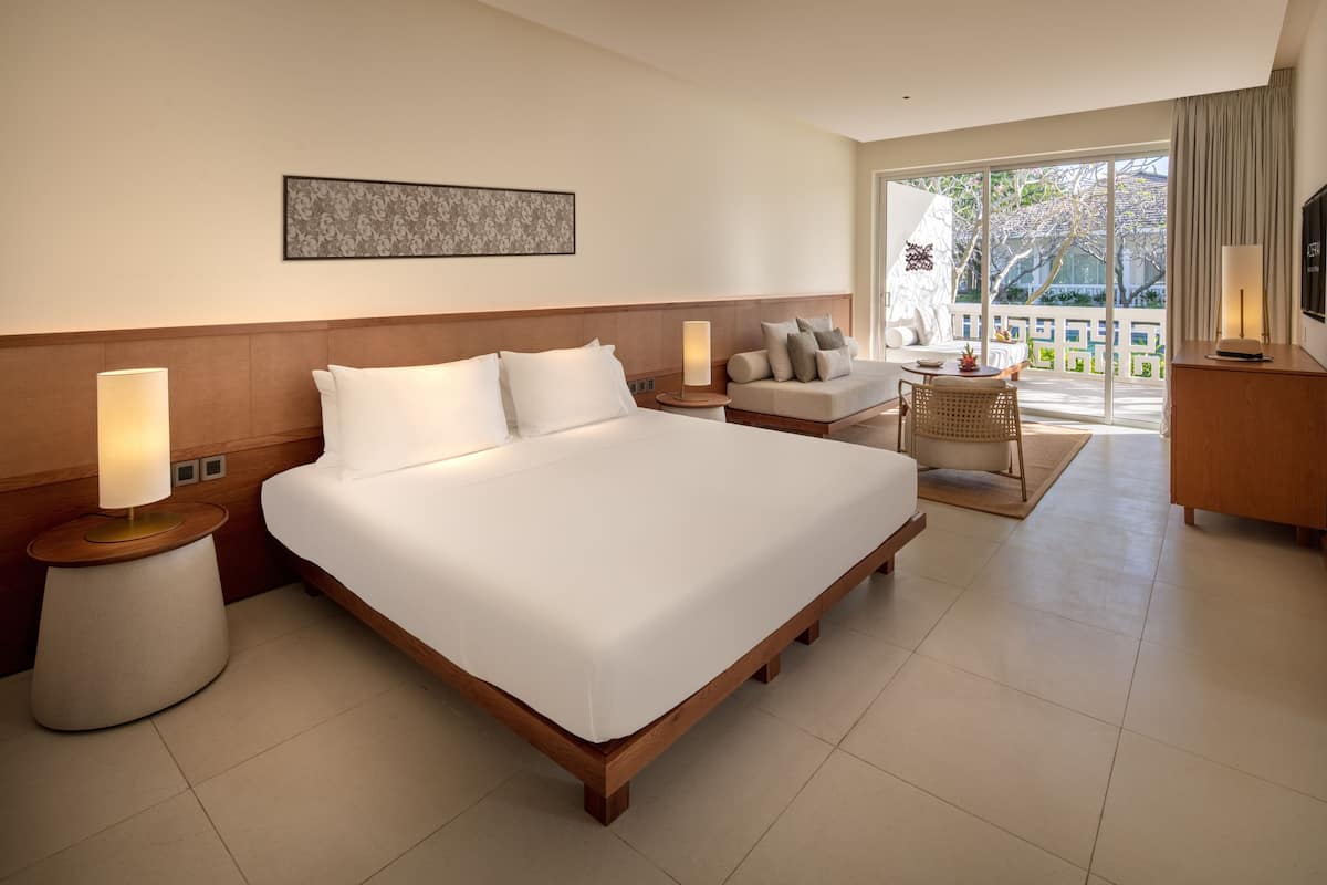 A modern hotel room with a large bed, a sofa, a TV, and a balcony with a view of trees.