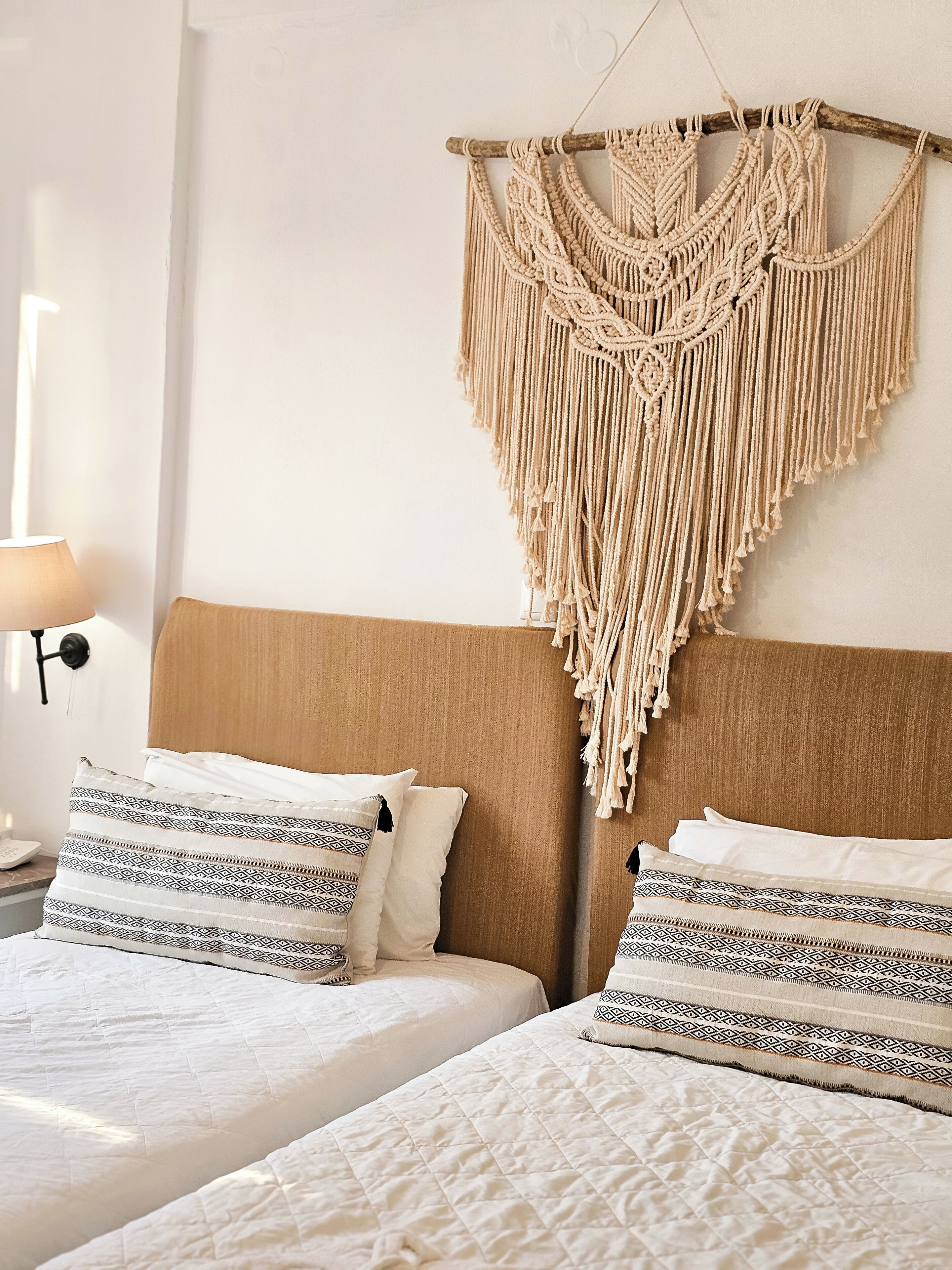 Een hotelkamer met twee bedden, een macramé wanddecoratie, gestreepte kussens en een bedlamp.