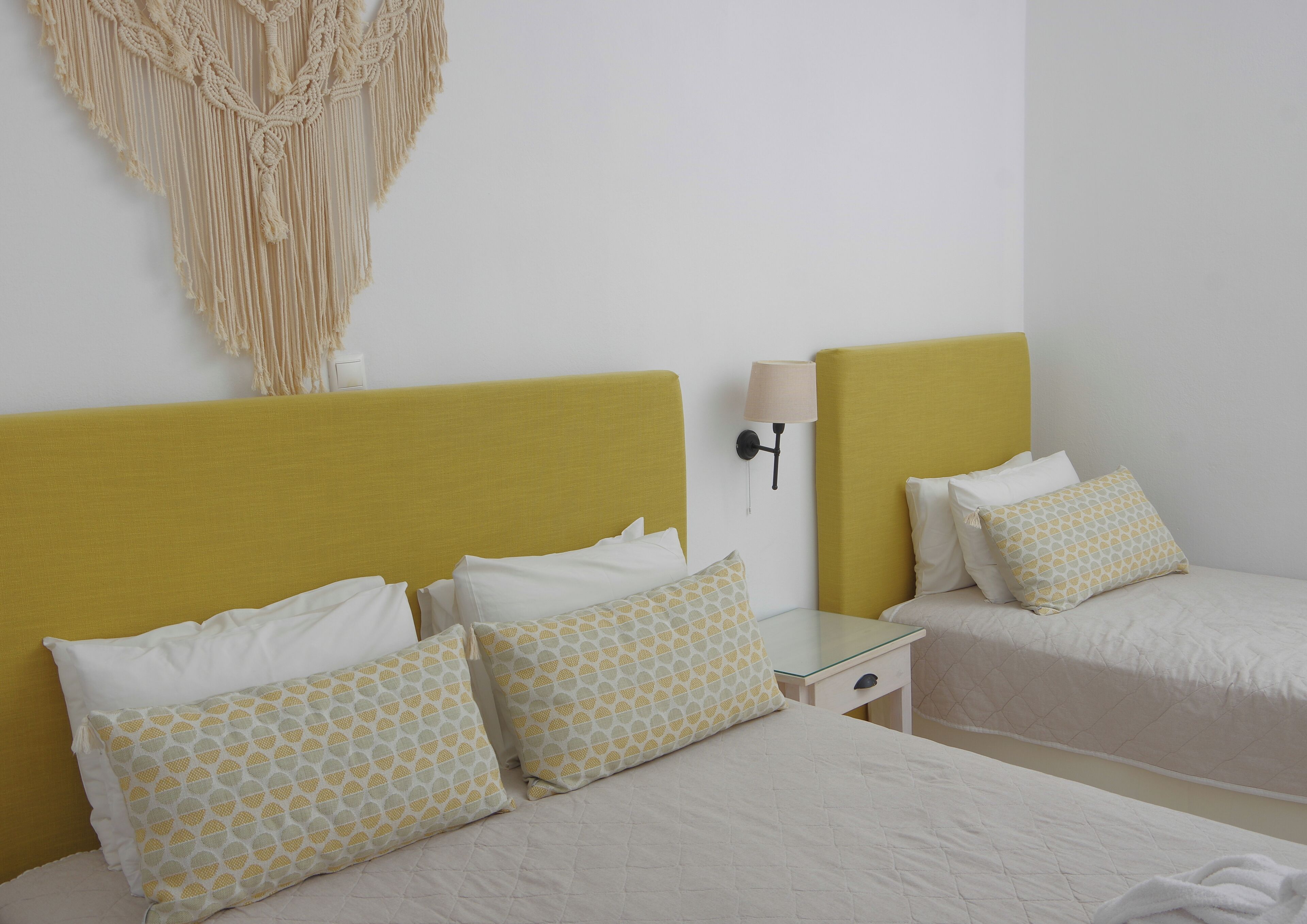 A hotel room with two beds, a bedside table, a lamp, and a macrame wall hanging.