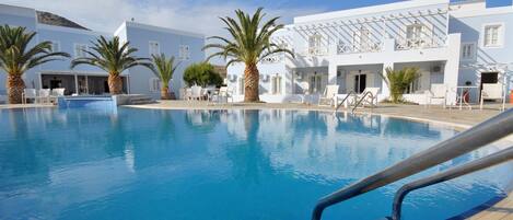Una piscina con sillas de descanso y palmeras, rodeada por un edificio blanco con balcones.