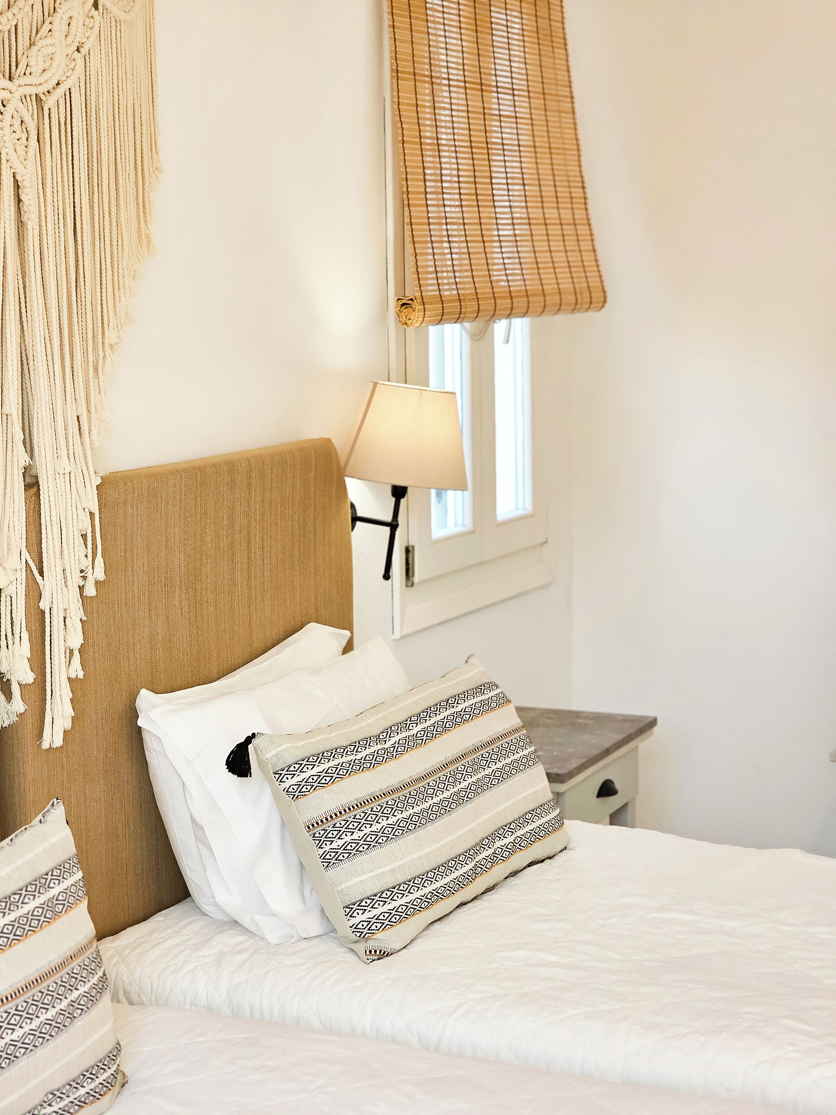 Una cama bien tendida con almohadones estampados, un colgador de macramé, una lámpara de noche y una ventana con persiana de bambú.