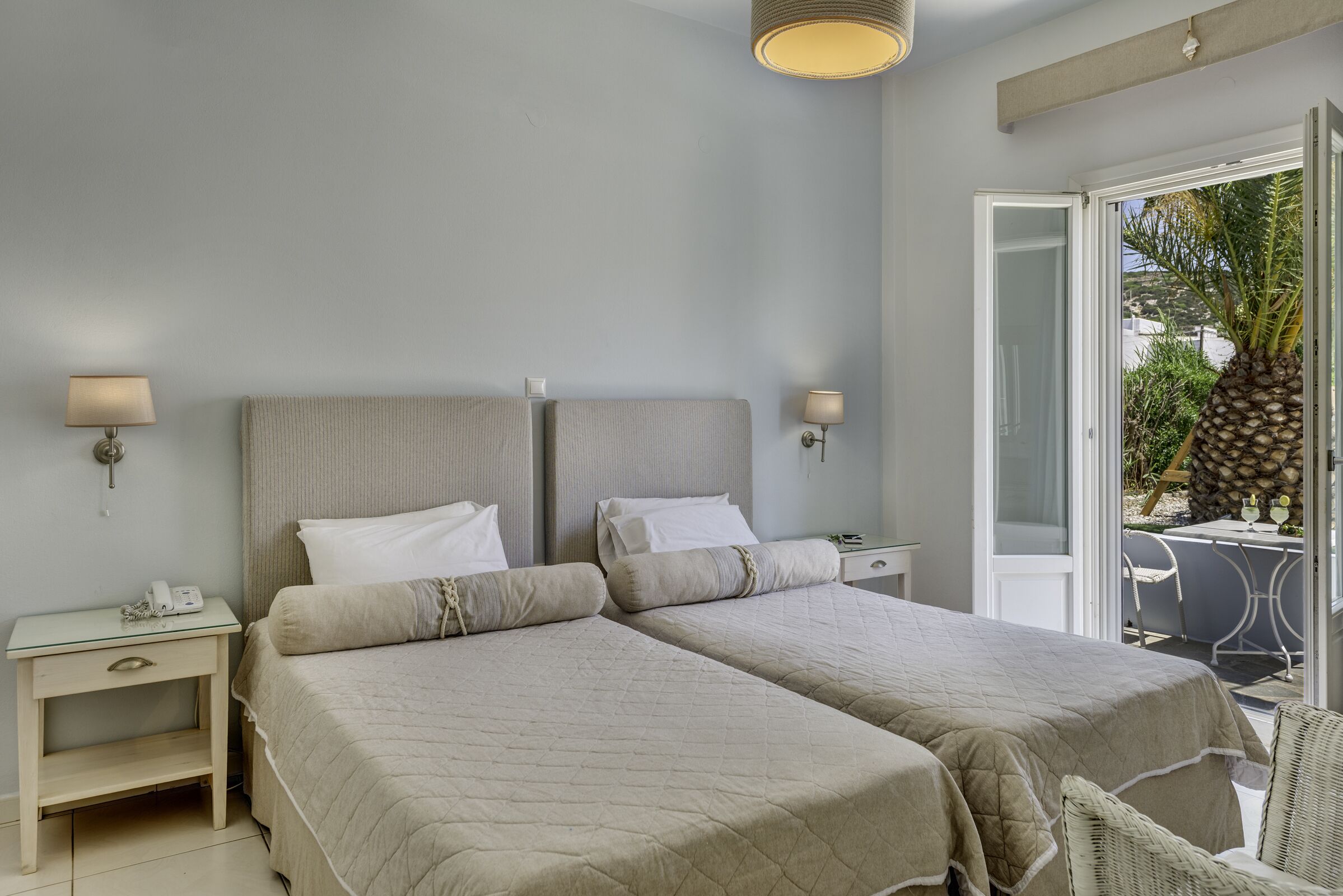 Cama doble con dos mesitas de noche, una lámpara y vista a una palmera a través de una puerta abierta.