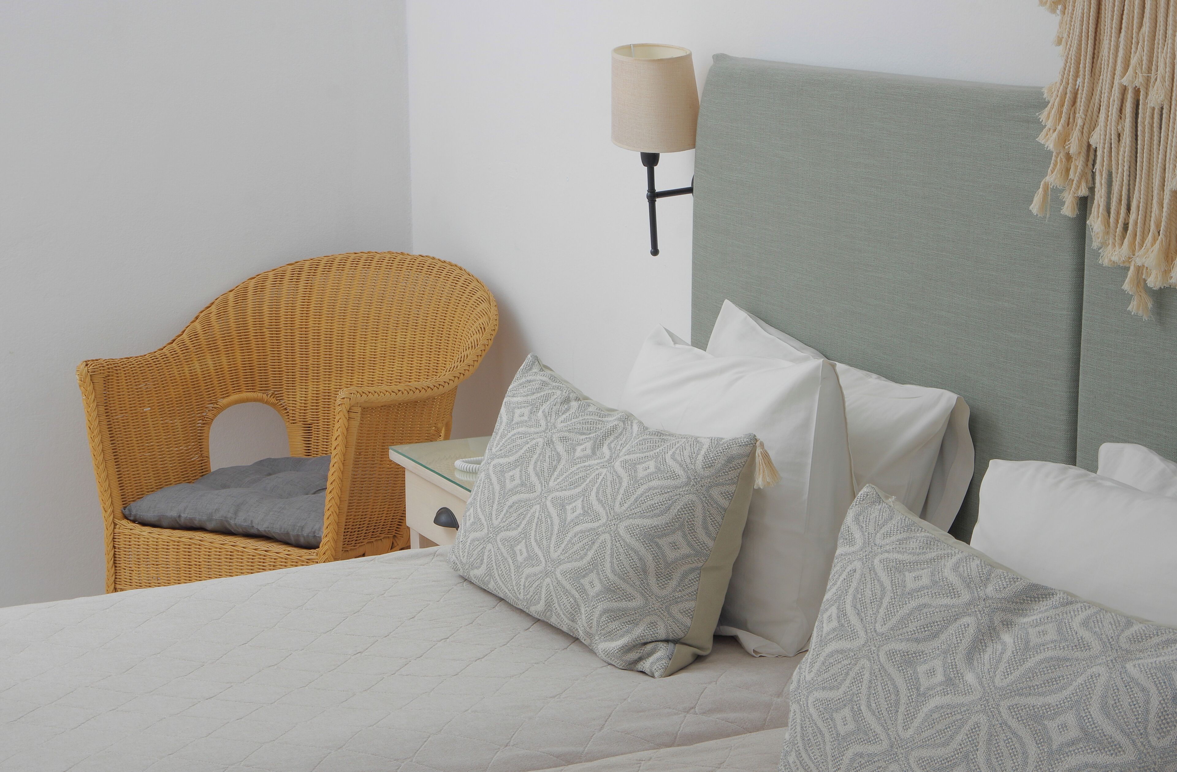 Un dormitorio ordenado con cama, almohadas, una silla de mimbre, una mesita de noche y una lámpara de pared.