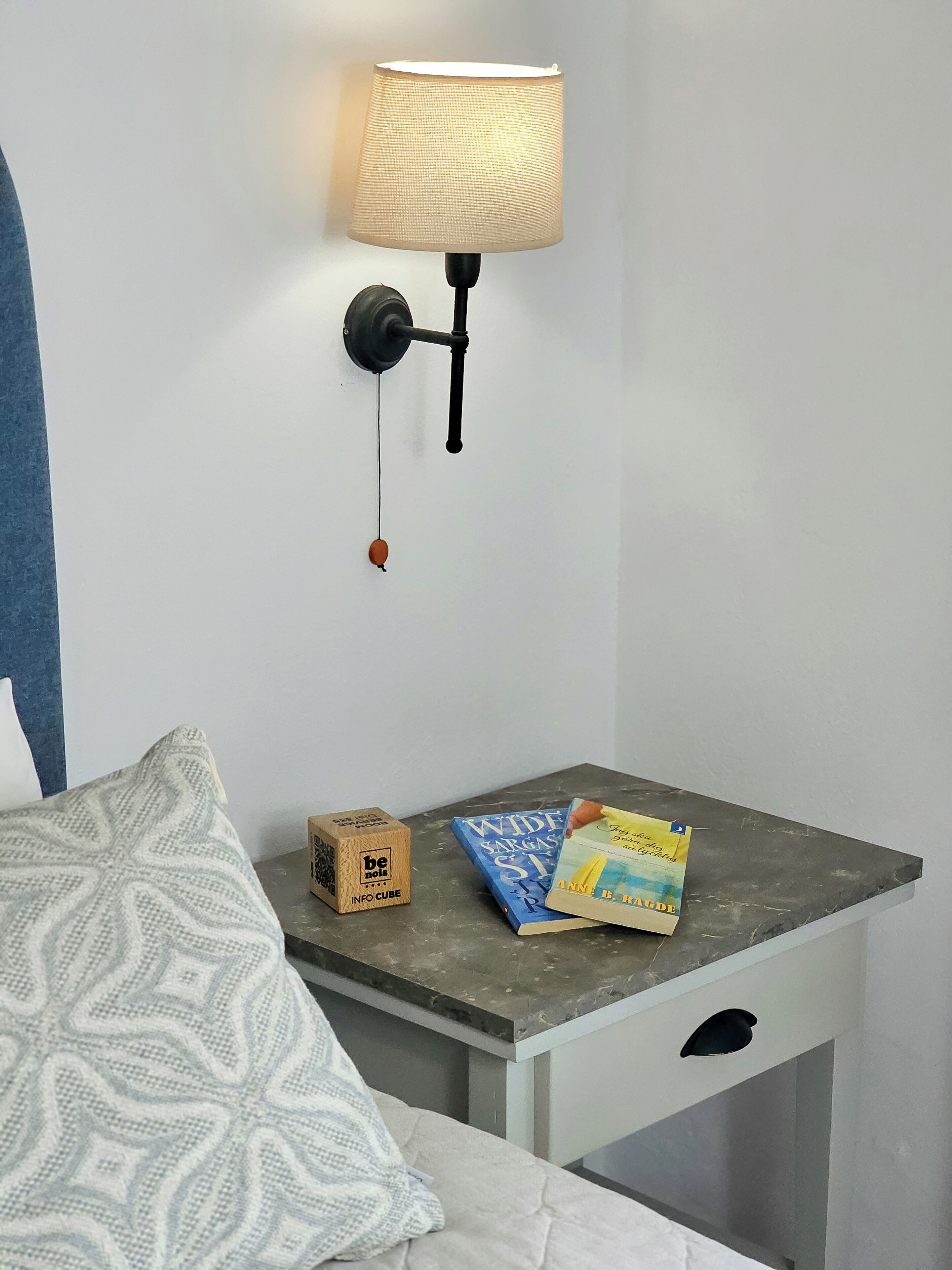 A bedroom with a nightstand, a lamp, a patterned pillow, a book, and a wooden box labeled "be nice".