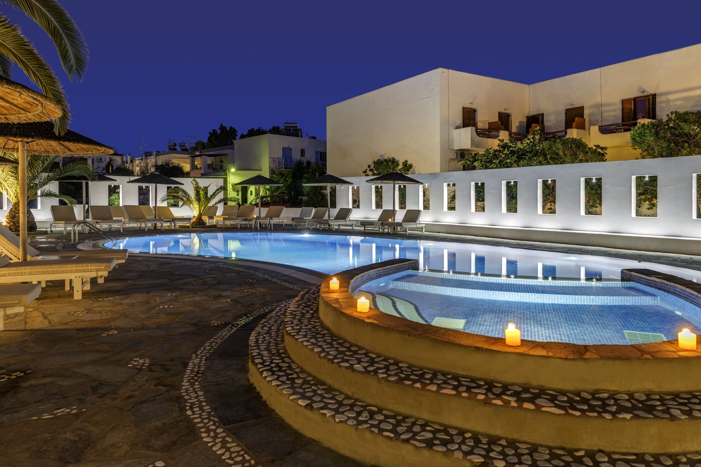 A pool area with lounge chairs and a hot tub, surrounded by a stone deck and a white building with windows.