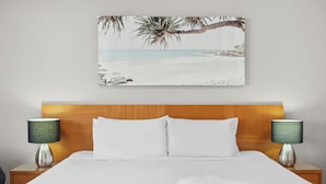Ein ordentlich bezogenes Bett mit weißen Leinen, zwei Nachttischlampen und ein Gemälde mit einer tropischen Strandansicht an der Wand.