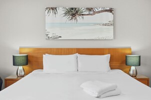 Cama bem arrumada com lençóis brancos, duas luminárias de cabeceira e uma pintura de uma paisagem praiana tropical na parede.