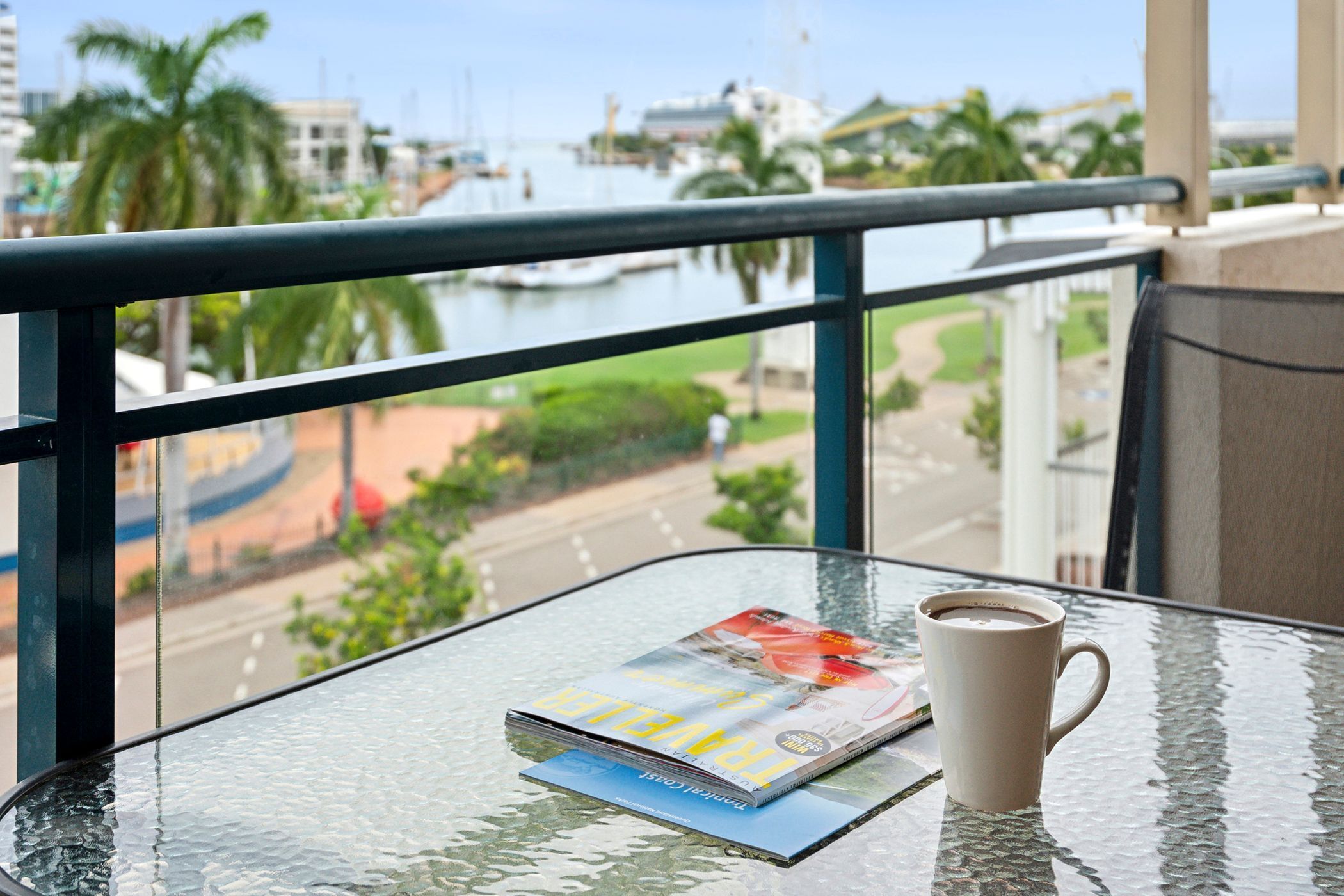 Ein Glastisch mit einer Zeitschrift, einer Tasse Kaffee und Blick auf einen Yachthafen mit Booten und Palmen.
