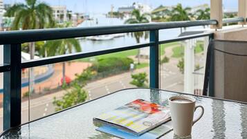 Ein Glastisch mit einer Zeitschrift, einer Tasse Kaffee und Blick auf einen Yachthafen mit Booten und Palmen.