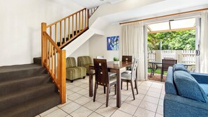 Ein geräumiger Wohnbereich mit einer Holztreppe, einem Esstisch mit Stühlen, einem Sofa und Blick auf einen Garten durch eine Glastür.
