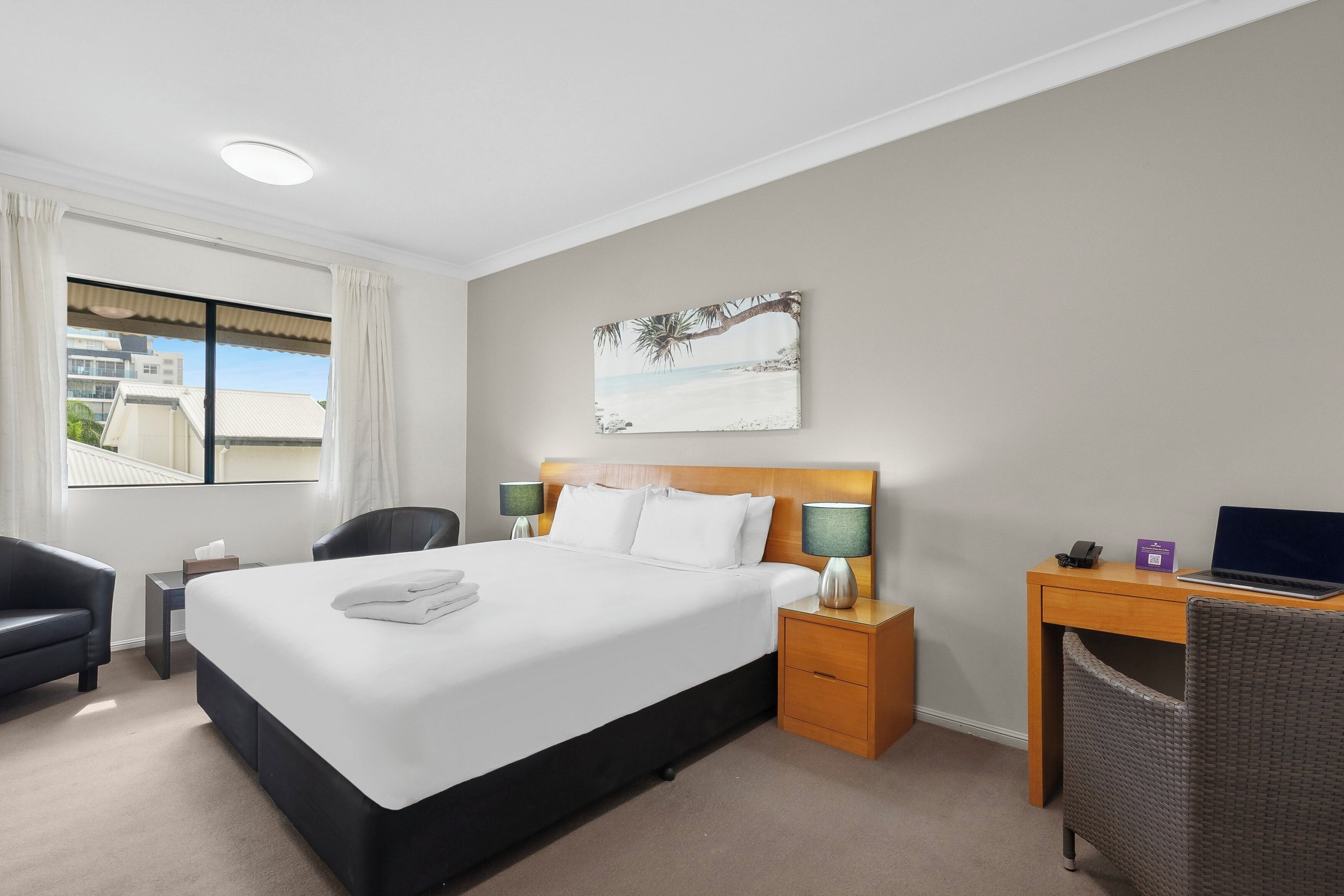 A hotel room with a large bed, a desk, two chairs, a small table with a lamp, and a painting on the wall.