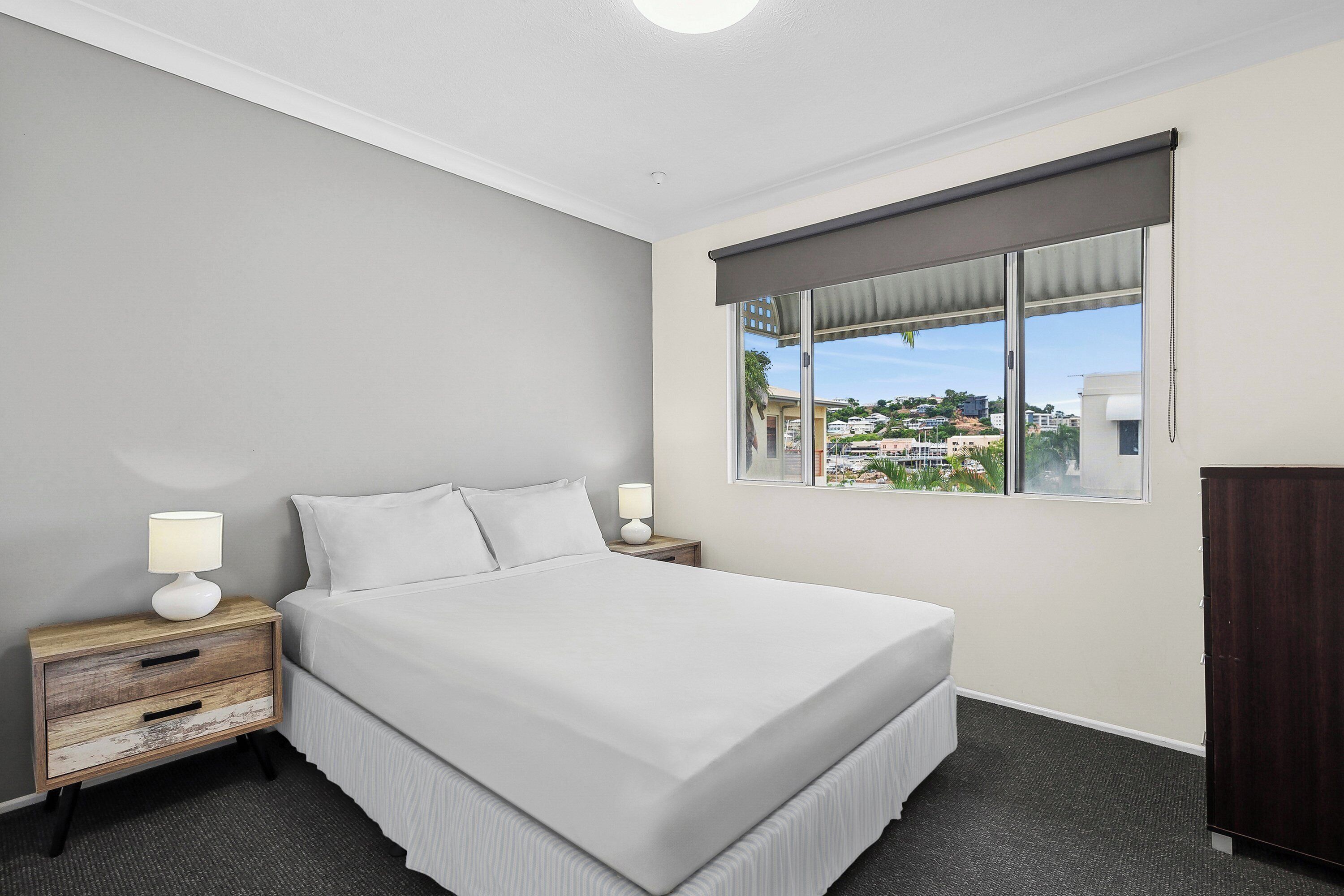 A hotel room with a large bed, a nightstand with a lamp, a wardrobe, and a window with a view of the city.