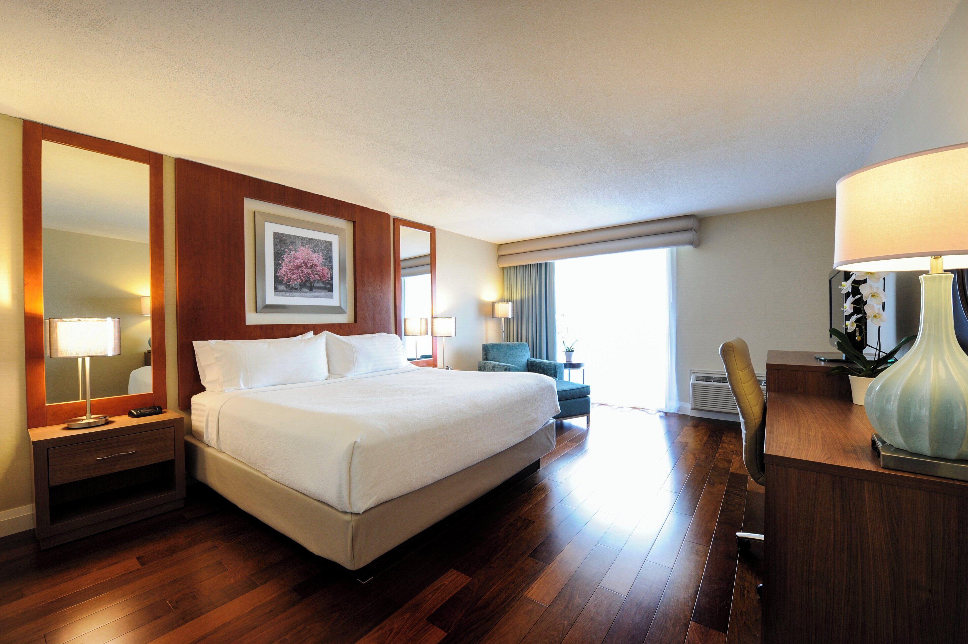 Une chambre d’hôtel avec un grand lit, un bureau, une chaise, une lampe de bureau, un miroir et une fenêtre avec des rideaux.