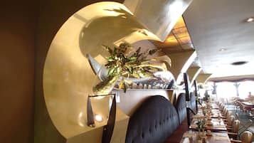 Interior de restaurante con una larga mesa de comedor, asientos acolchados y un centro de mesa decorativo con planta.
