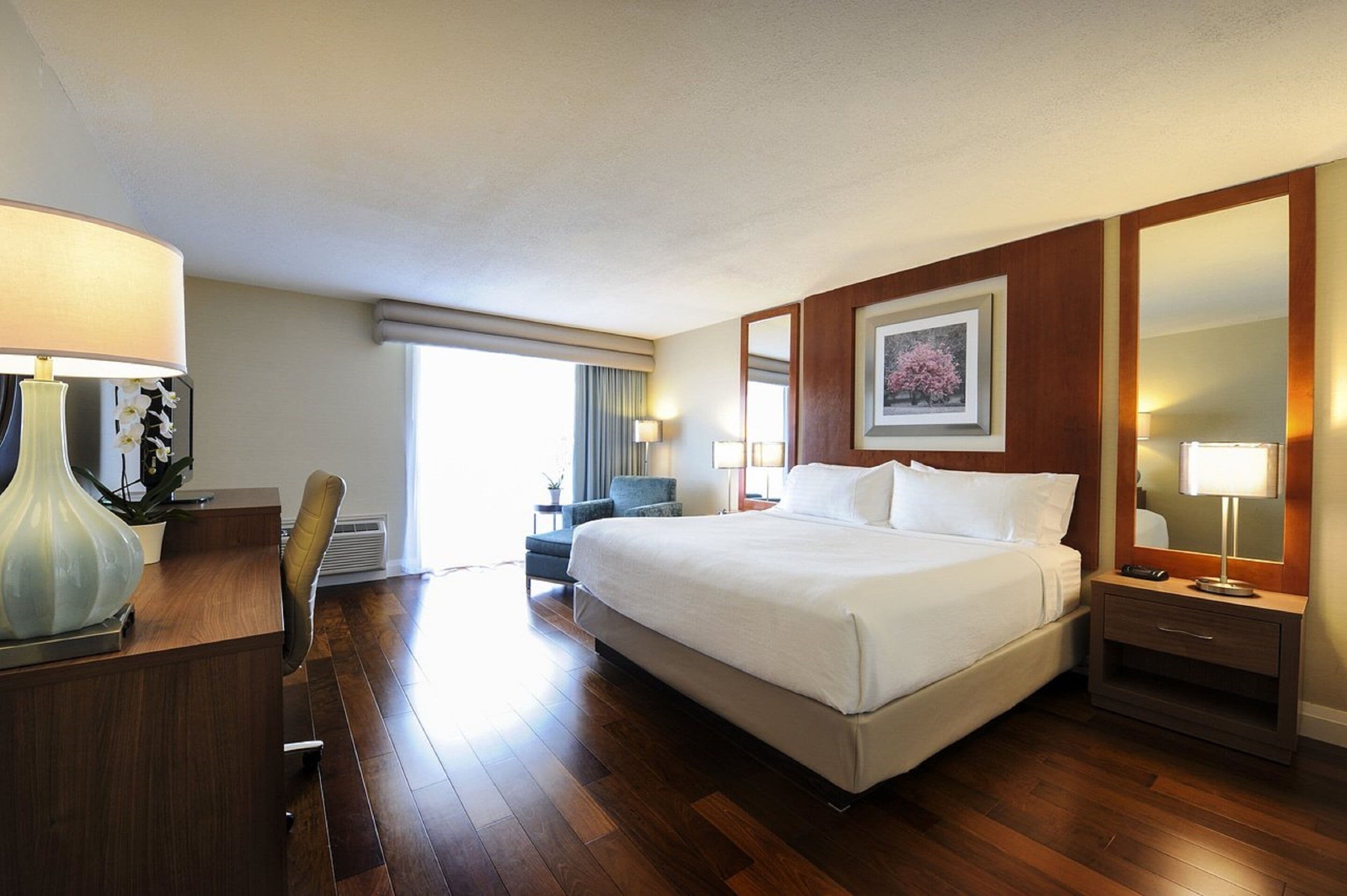 A hotel room with a large bed, a desk with a lamp, a chair, a bedside table with a lamp, a framed picture on the wall, and a window with curtains.