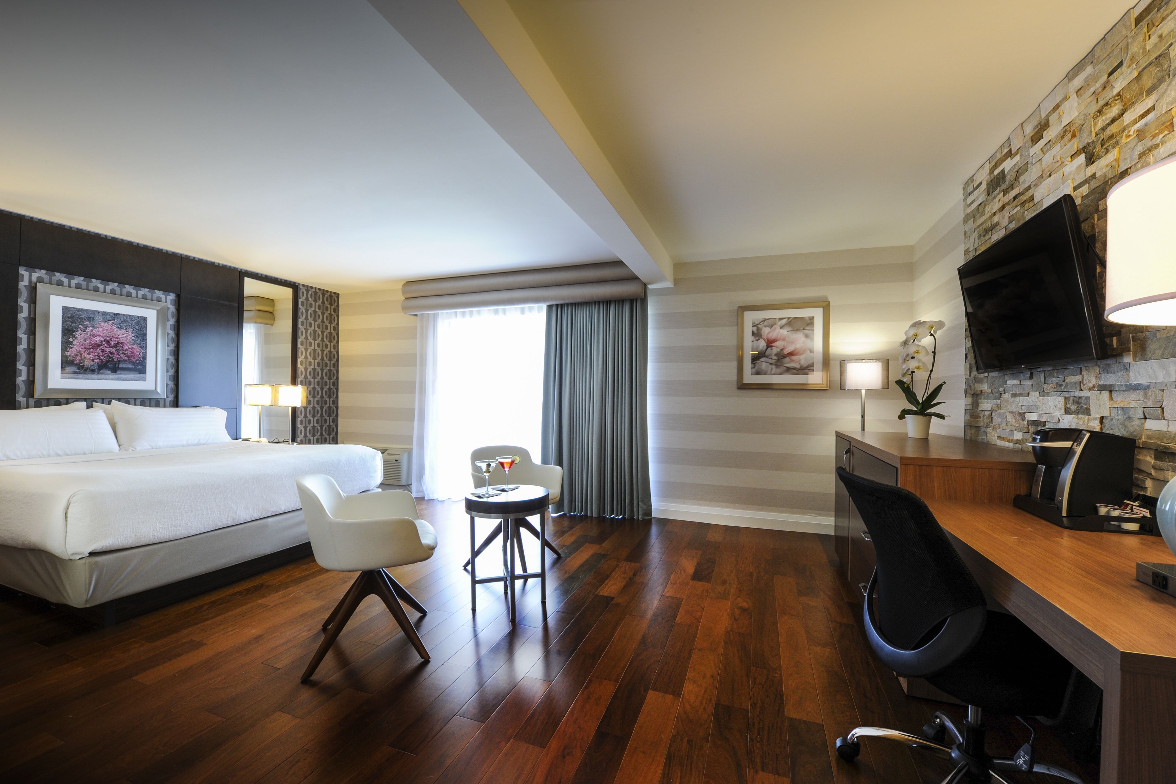 A hotel room with a bed, two chairs, a desk, a TV, and a window with curtains.