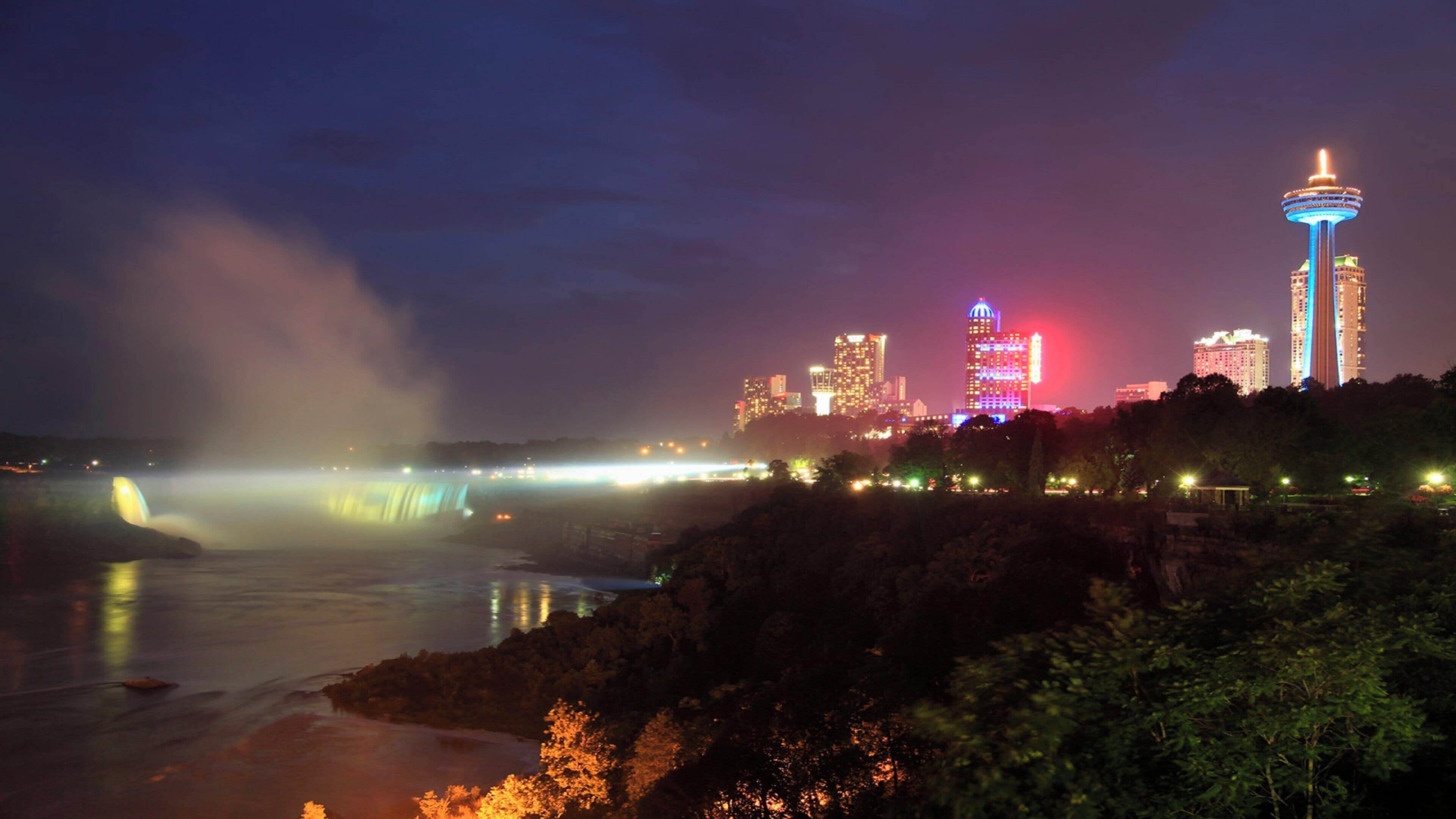 A nighttime cityscape with a prominent tower and a waterfall.