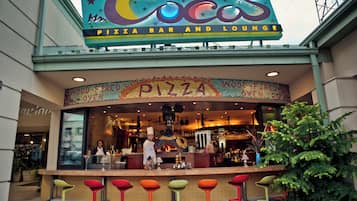 Un bar-pizzeria avec un espace détente, une enseigne colorée, un four à bois et une rangée de tabourets aux couleurs vives.