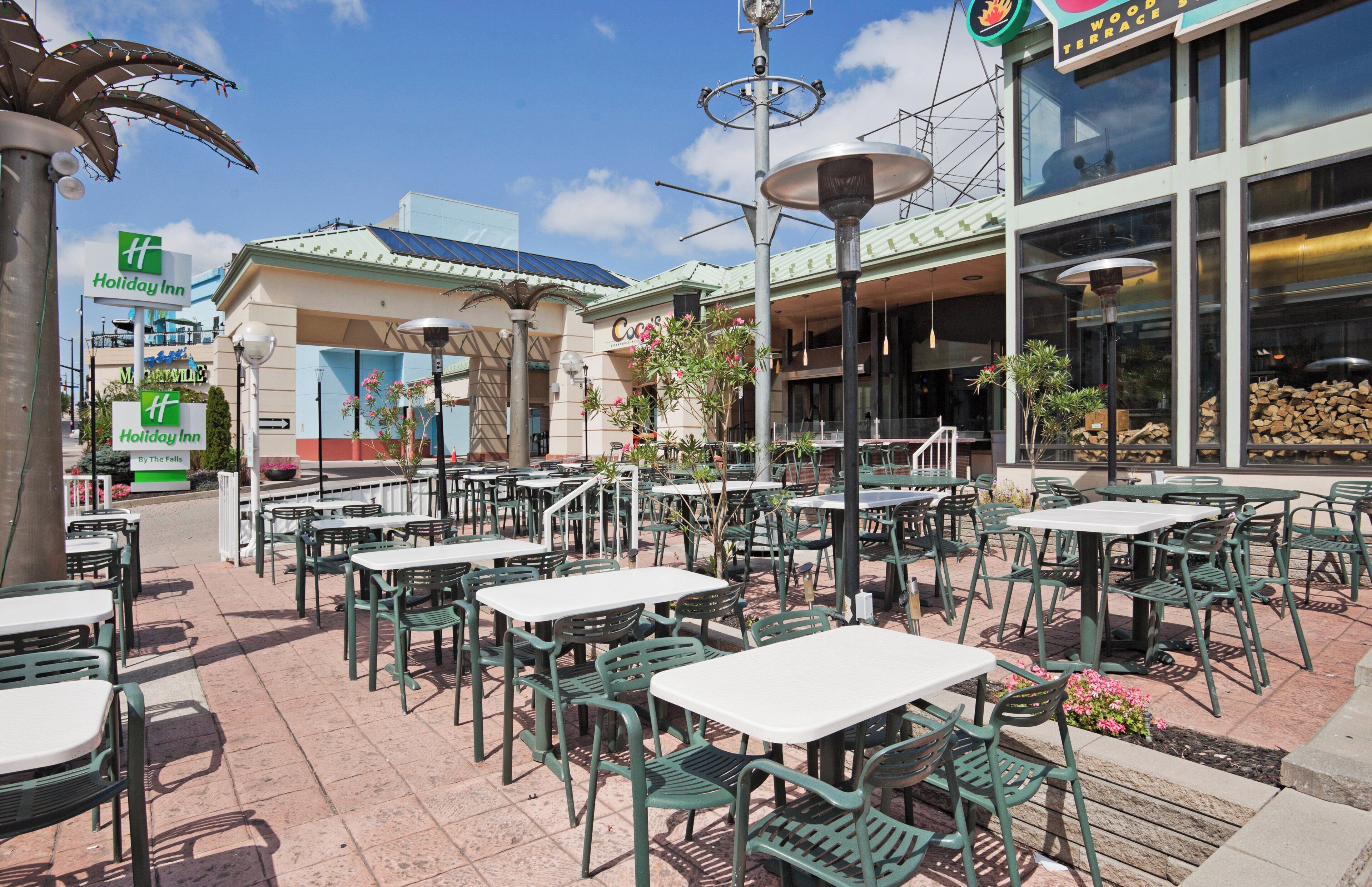 Un espace extérieur aménagé en terrasse, avec plusieurs tables et chaises, un bâtiment portant une enseigne « Holiday Inn » et un restaurant appelé « Coco’s ».