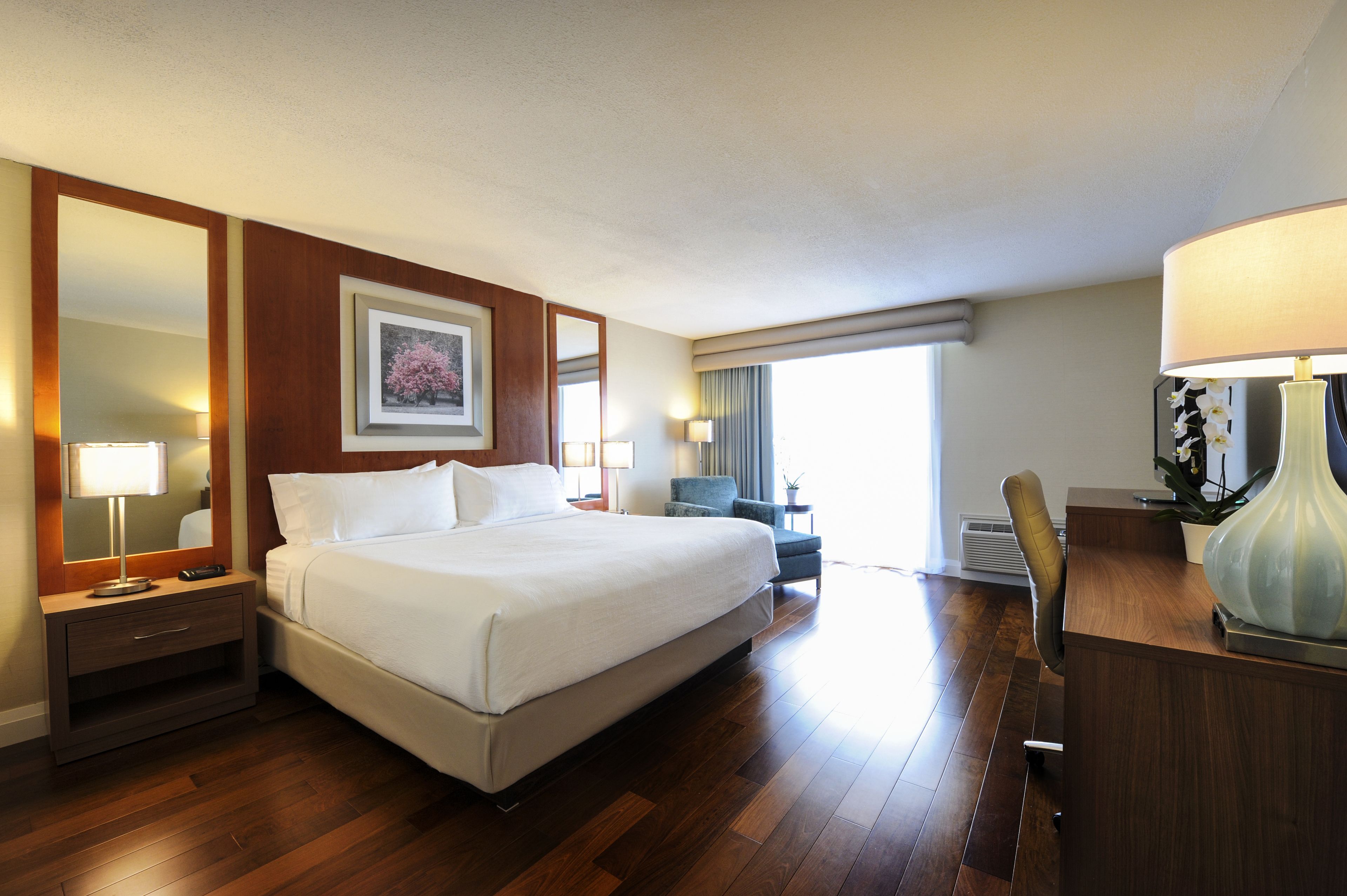 Quarto de hotel com uma cama grande, uma escrivaninha, uma cadeira, um espelho e uma janela com cortinas.