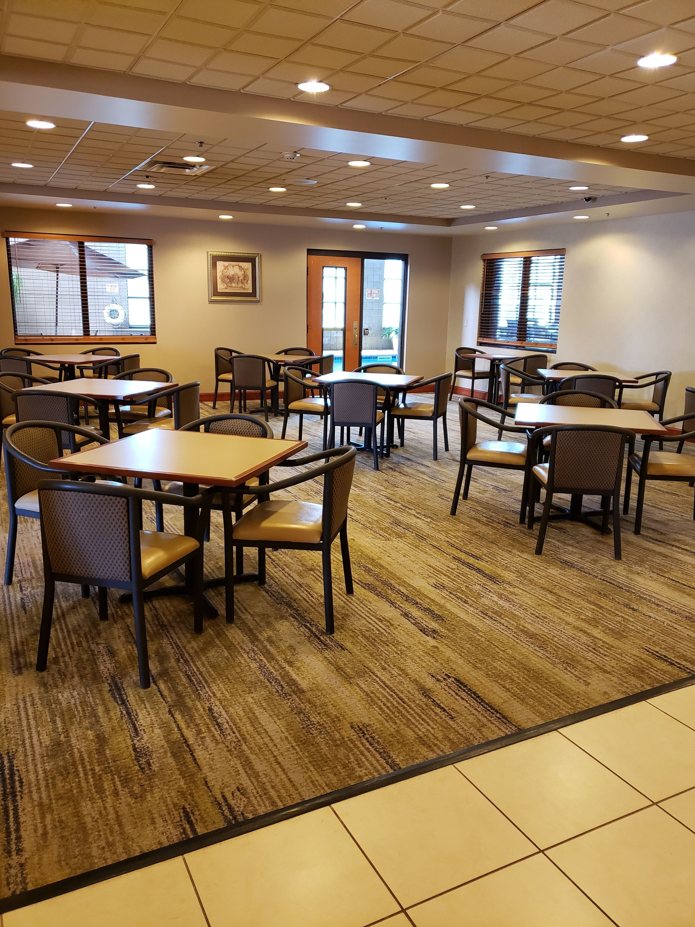 A hotel dining area with multiple tables and chairs, large windows with blinds, and a ceiling with recessed lighting.