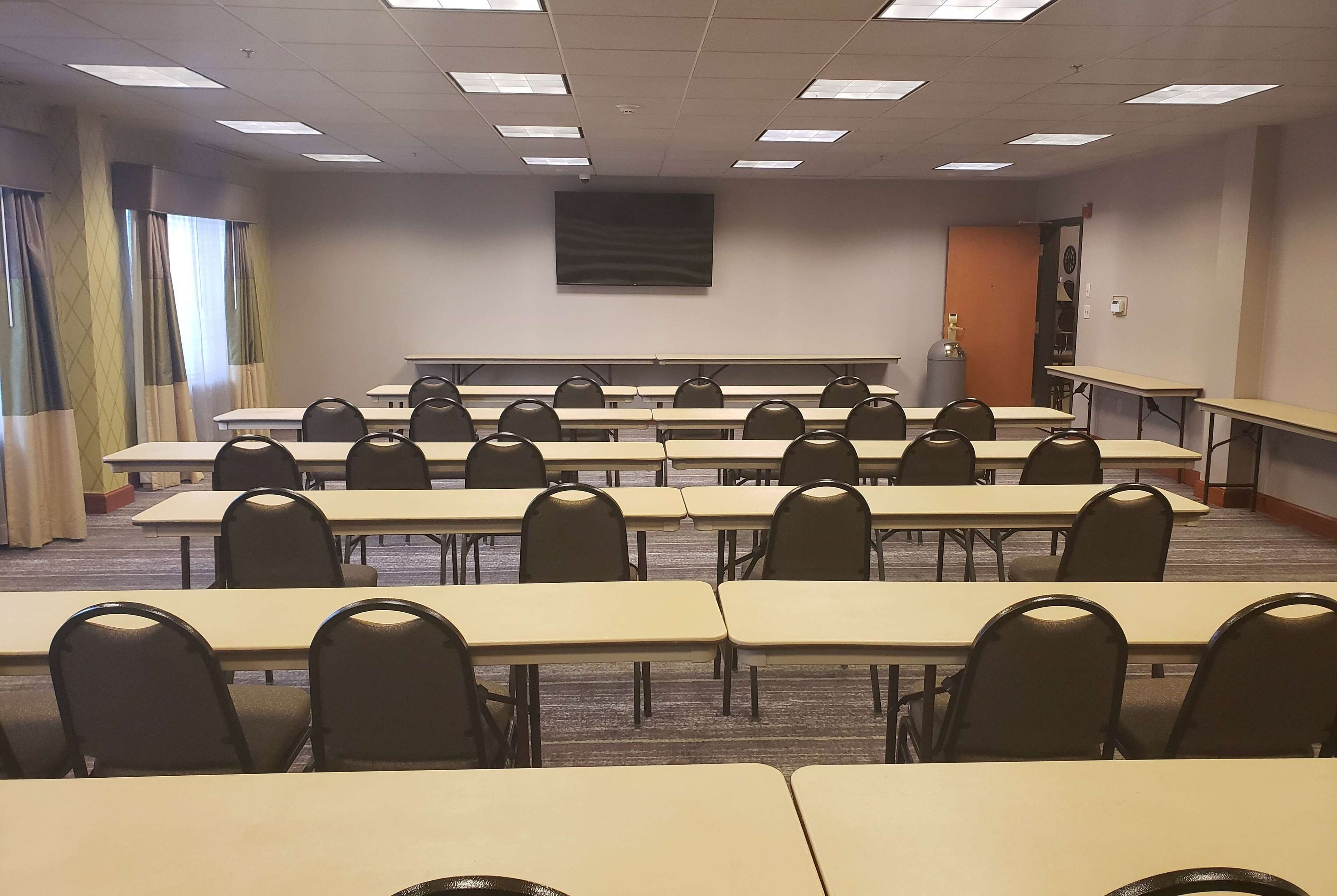 Un salón de conferencias con filas de mesas y sillas, una pantalla grande en la pared y una puerta al fondo.