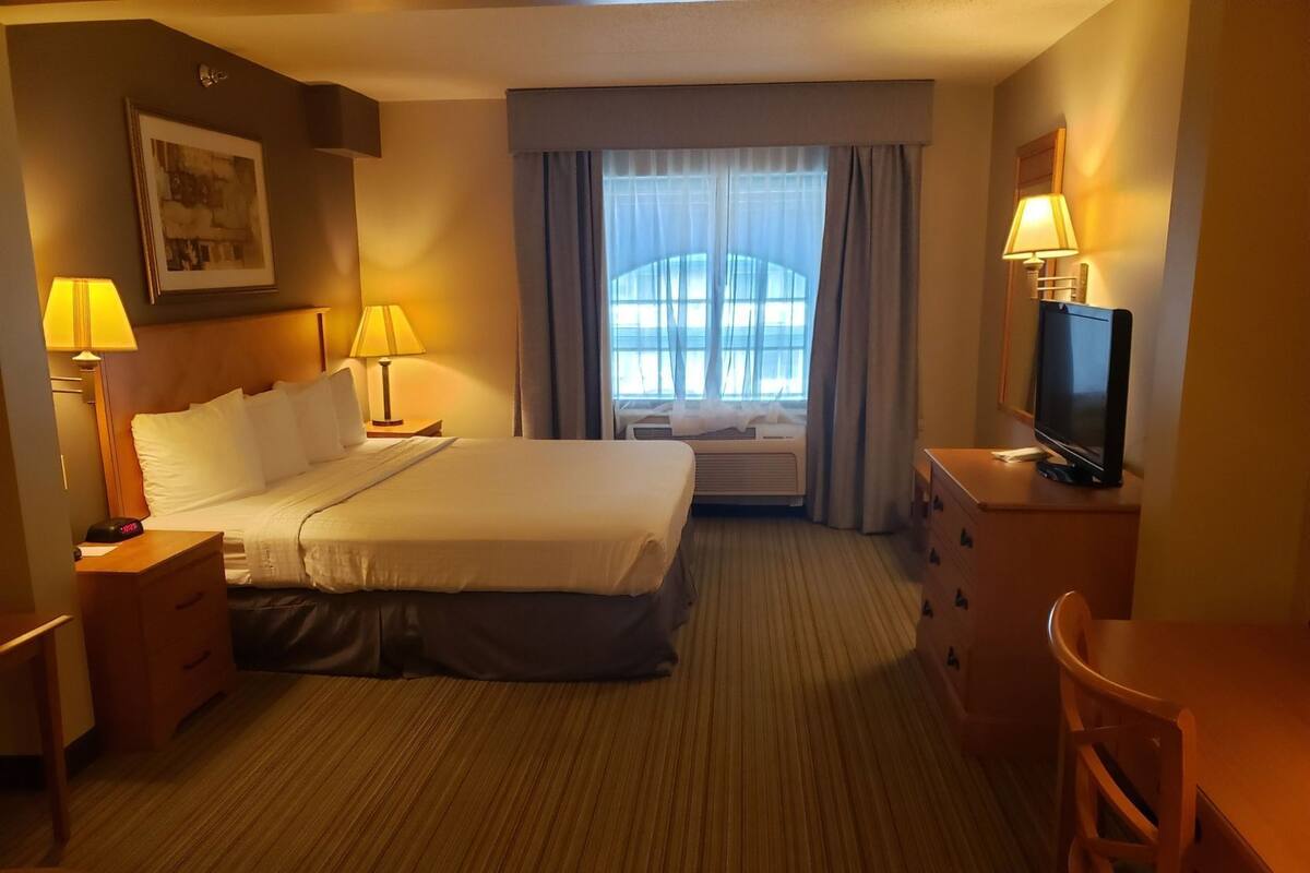 A hotel room with a bed, bedside lamps, a desk with a TV, a chair, a dresser, and a window with curtains.