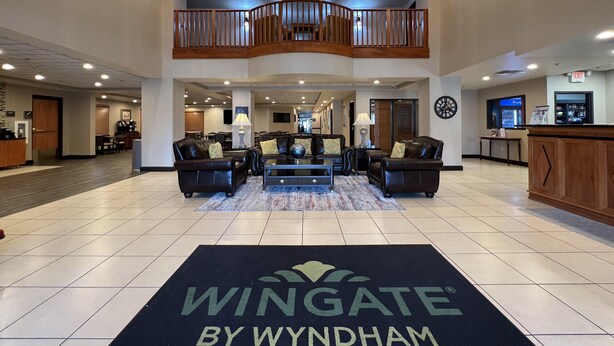 A spacious lobby with a high ceiling, a central seating area with leather sofas, a staircase to an upper level, and a reception desk with a clock and a digital screen. - Wingate by Wyndham Coon Rapids (Coon Rapids)