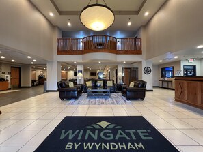 A spacious lobby with a high ceiling, a central seating area with leather sofas, a staircase to an upper level, and a reception desk with a clock and a digital screen. - Wingate by Wyndham Coon Rapids (Coon Rapids)