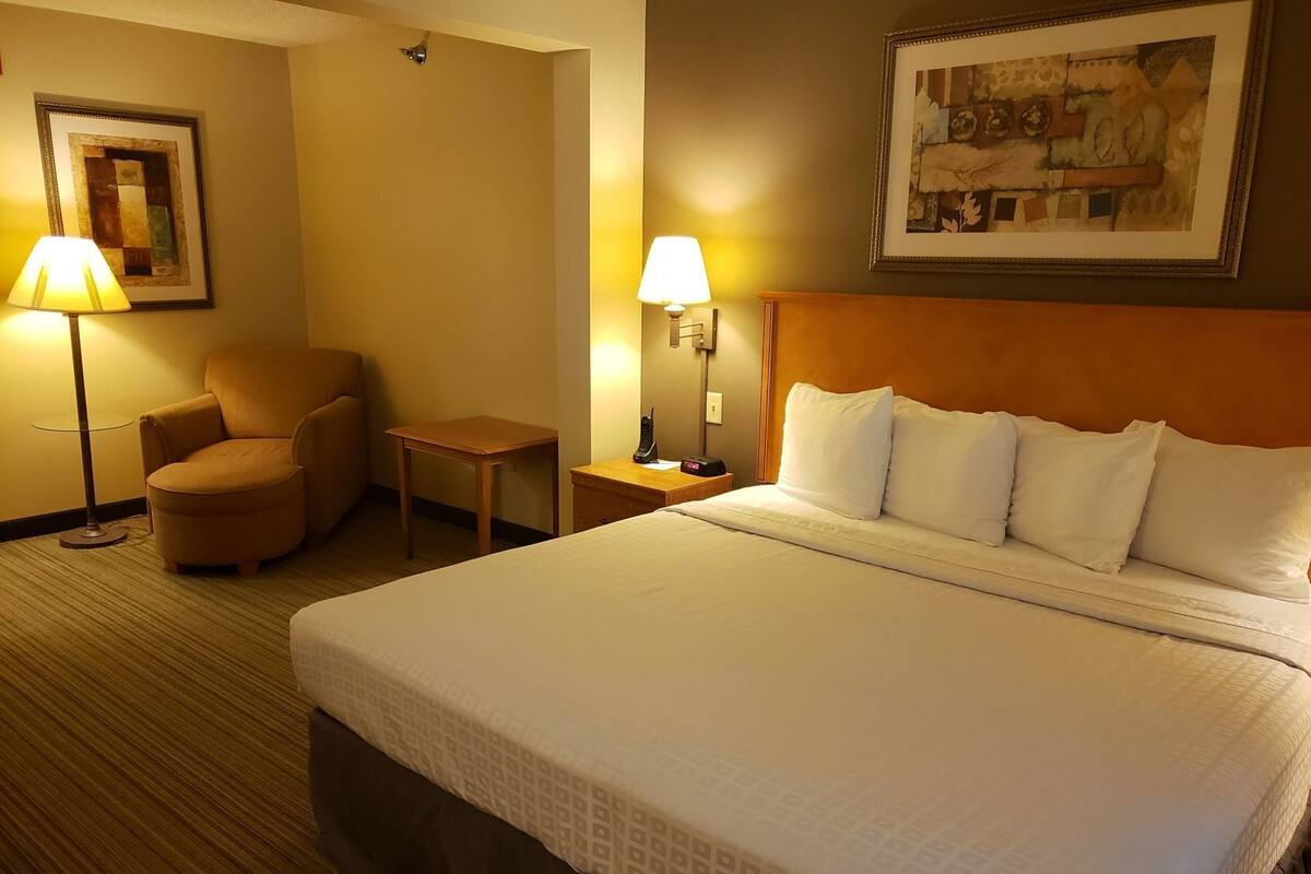 A hotel room with a bed, a chair, a small table, two lamps, and a painting on the wall.