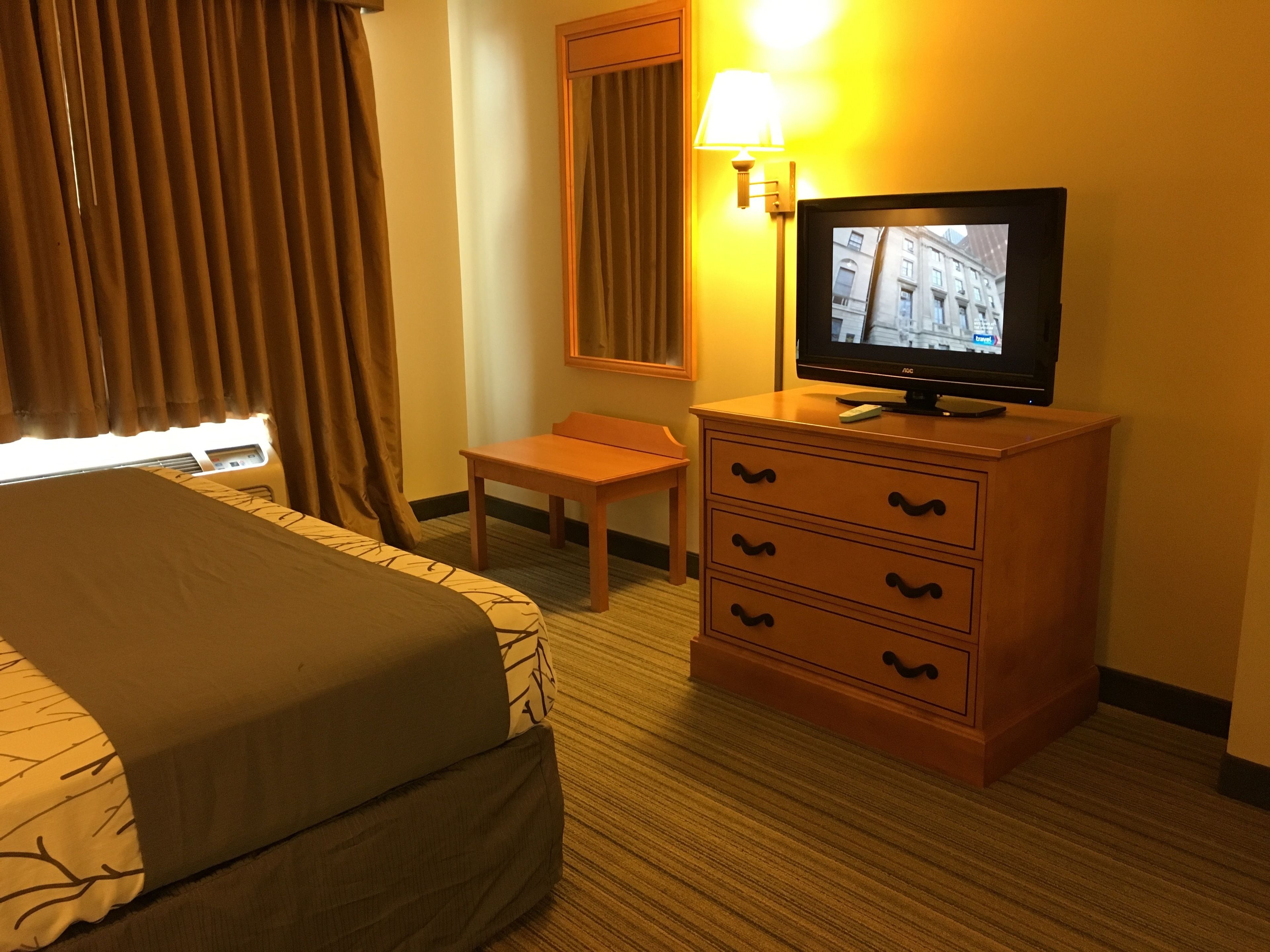 A hotel room with a bed, a wooden dresser with drawers, a television on the dresser, a lamp, and a window with curtains.