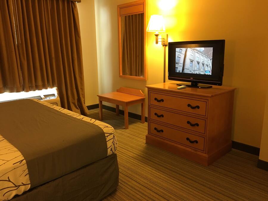 Habitación de hotel con cama, cómoda de madera con cajones, televisor sobre la cómoda, lámpara y ventana con cortinas.