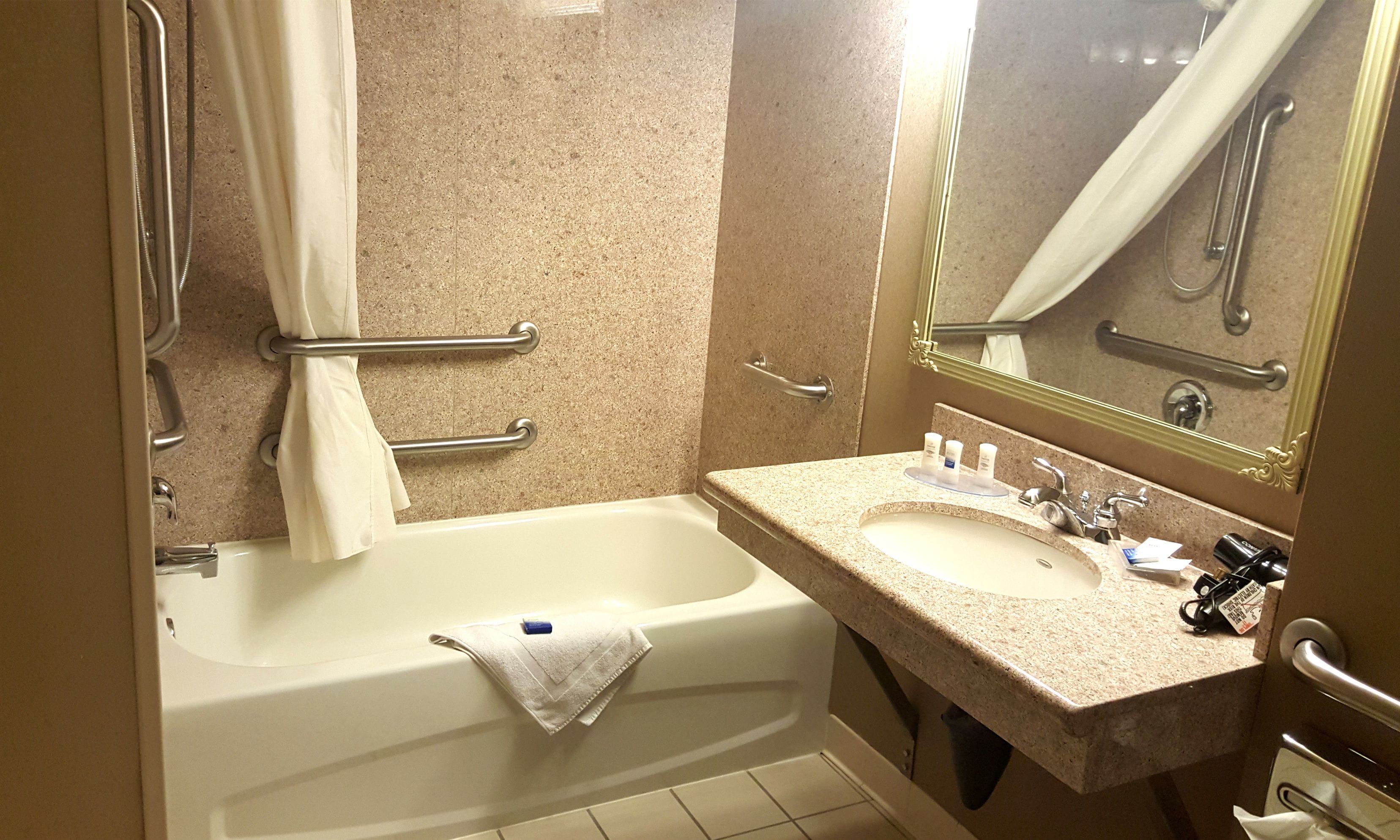A bathroom with a bathtub, a sink, and a mirror. There are handrails and a towel rack.