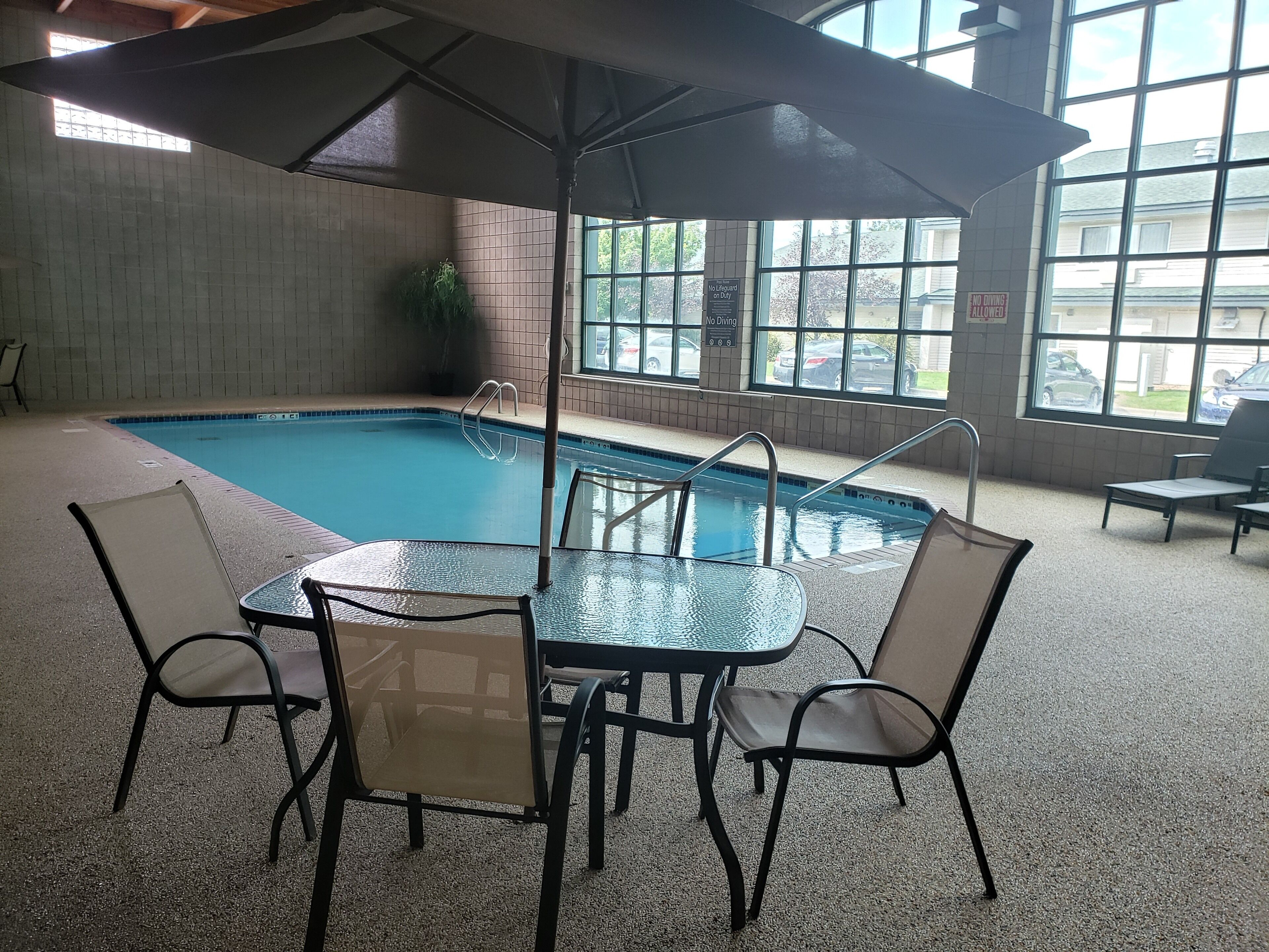 Una piscina interior con mesa y sillas de vidrio, rodeada de ventanas y vistas a coches aparcados afuera.