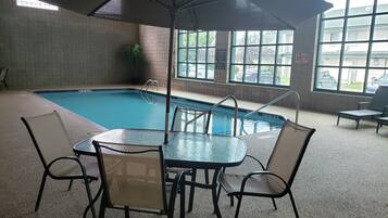 Una piscina interior con mesa y sillas de vidrio, rodeada de ventanas y vistas a coches aparcados afuera.