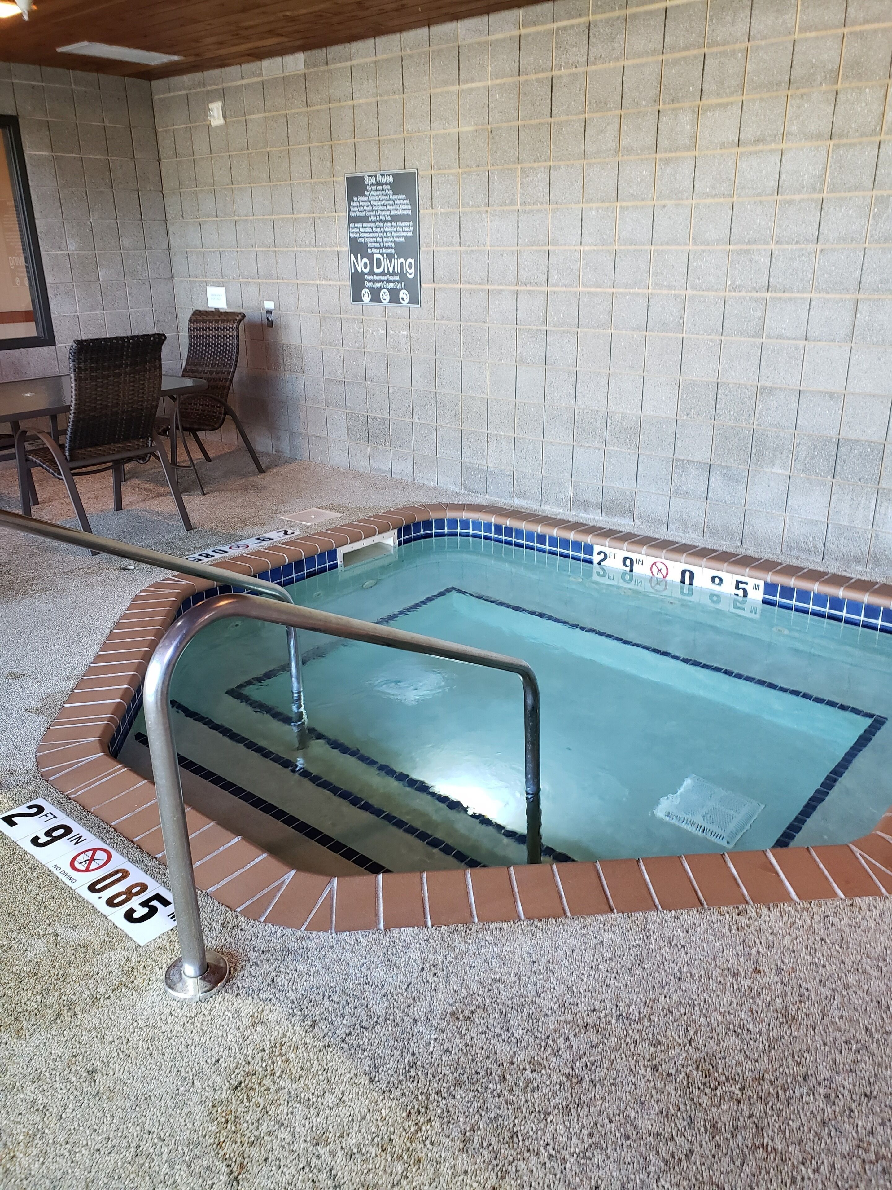 An indoor swimming pool with a tiled wall, a sign prohibiting diving, and a seating area with chairs.