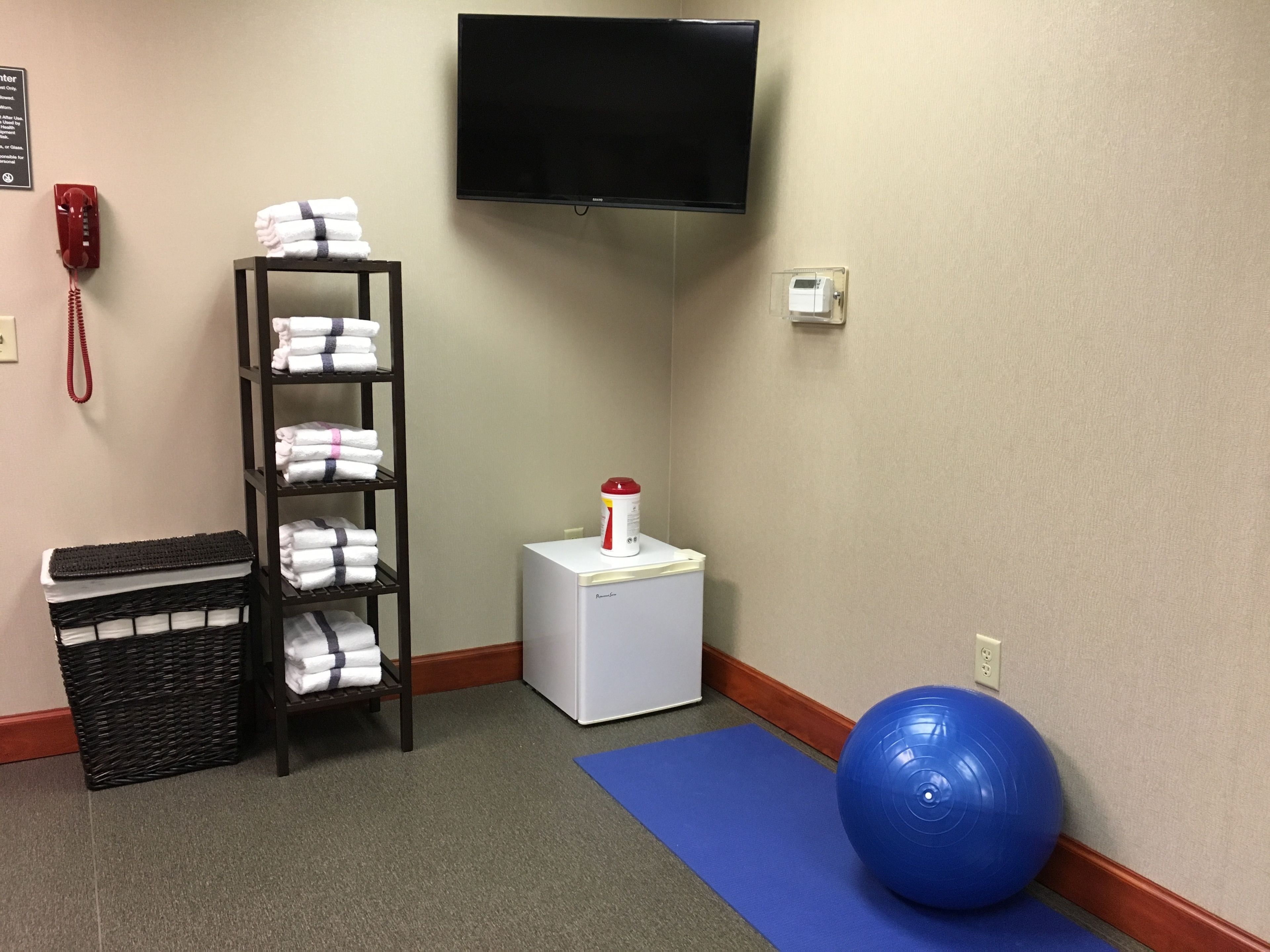 A corner room with a TV, a small white appliance, a shelf with towels, a wicker basket, and a blue exercise ball.