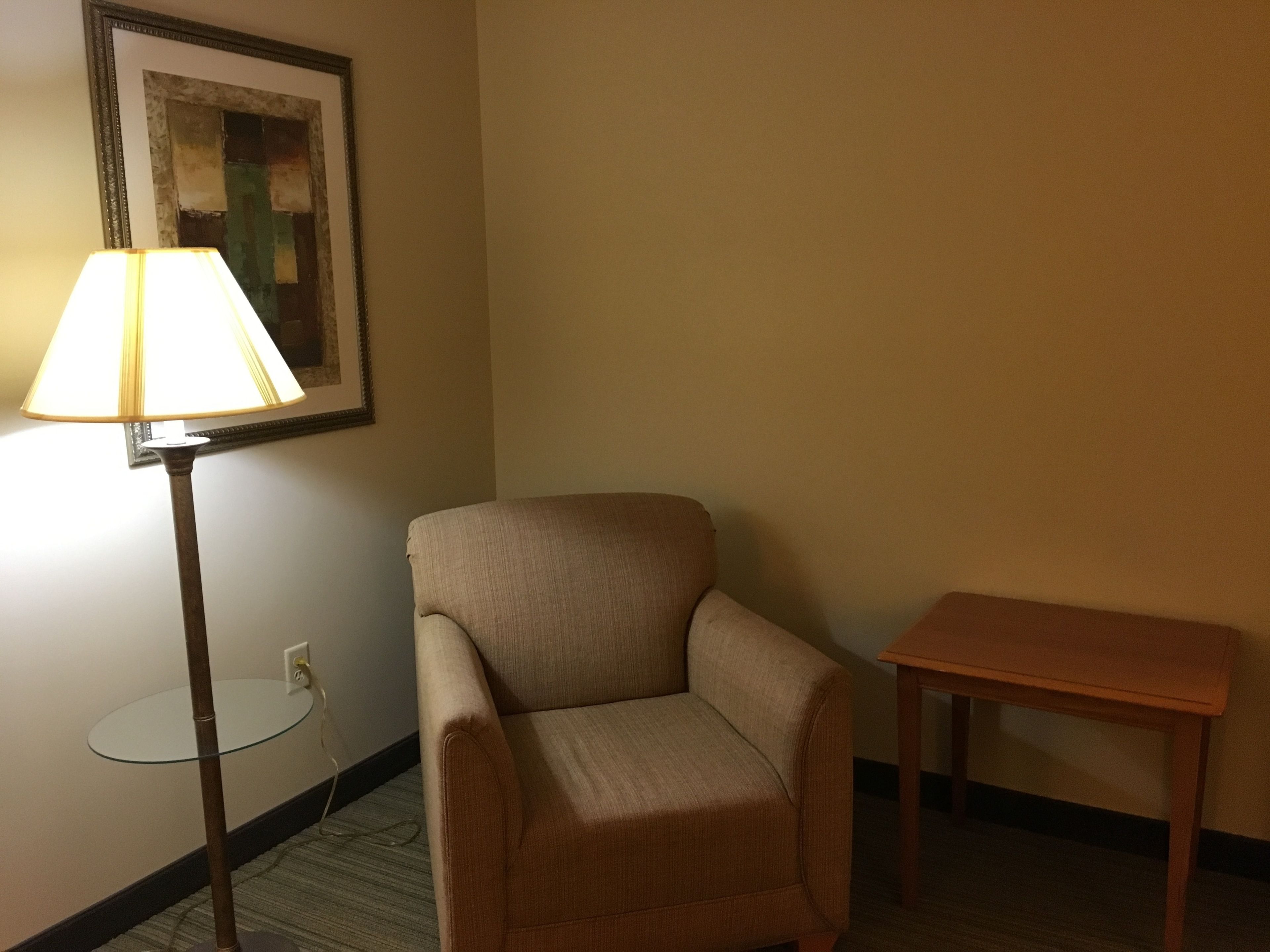 Una habitación con un sillón color beige, una pequeña mesa de madera, una lámpara de pie y una obra enmarcada en la pared.