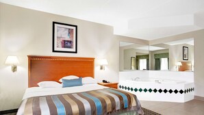 Habitación de hotel con una cama grande, cabecero de madera, dos lámparas de noche, un espejo amplio, baño con bañera e imagen enmarcadas en la pared.