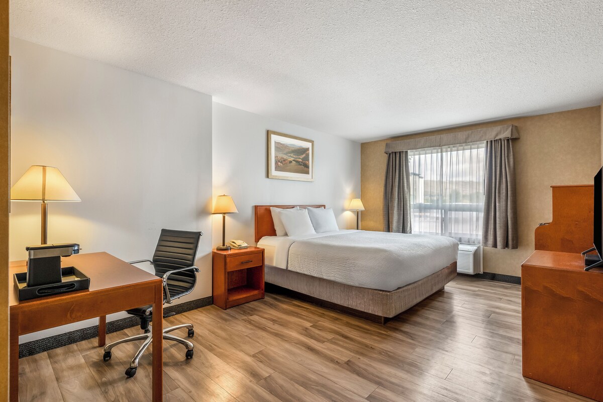 A hotel room with a bed, desk, chair, lamps, and a painting on the wall.