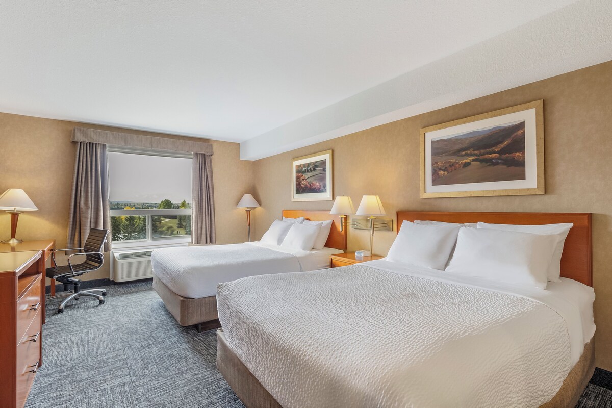A hotel room with two beds, a desk, a chair, a window with a view, and two wall-mounted lamps.