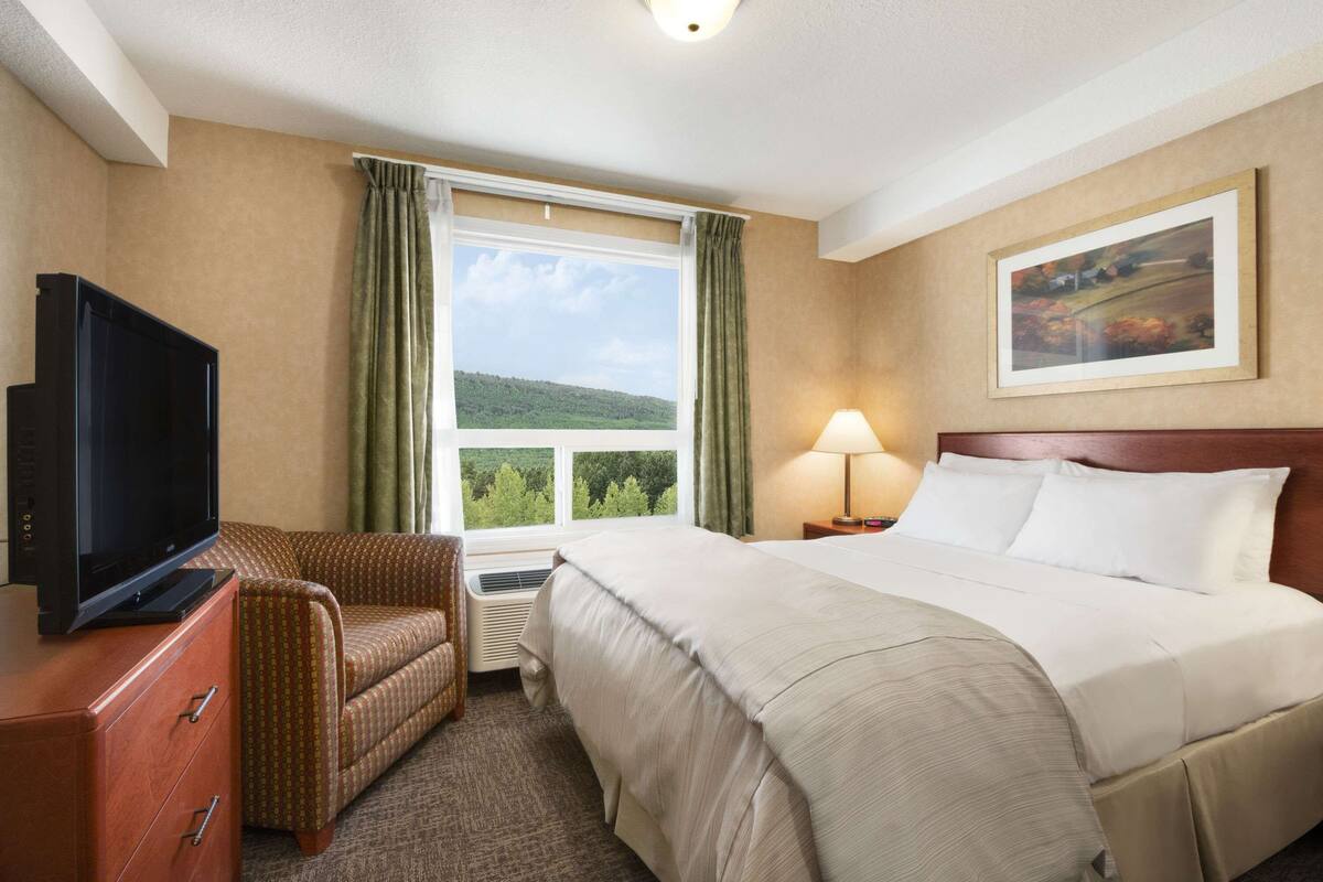 Una habitación de hotel con cama, televisor sobre una cómoda, una silla, ventana con cortinas, lámpara y un cuadro en la pared.