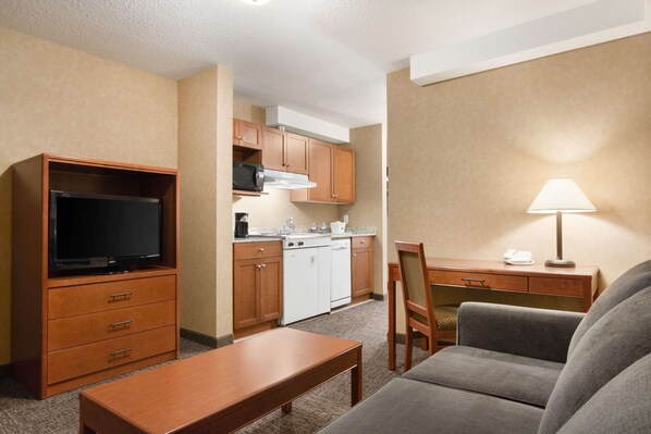 A compact hotel room with a kitchenette, a flat-screen TV, a sofa, a dining table, and a lamp.