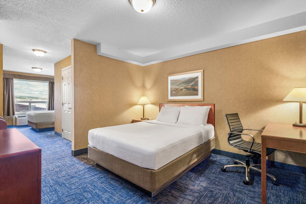A hotel room with a bed, desk, chair, and a painting on the wall.