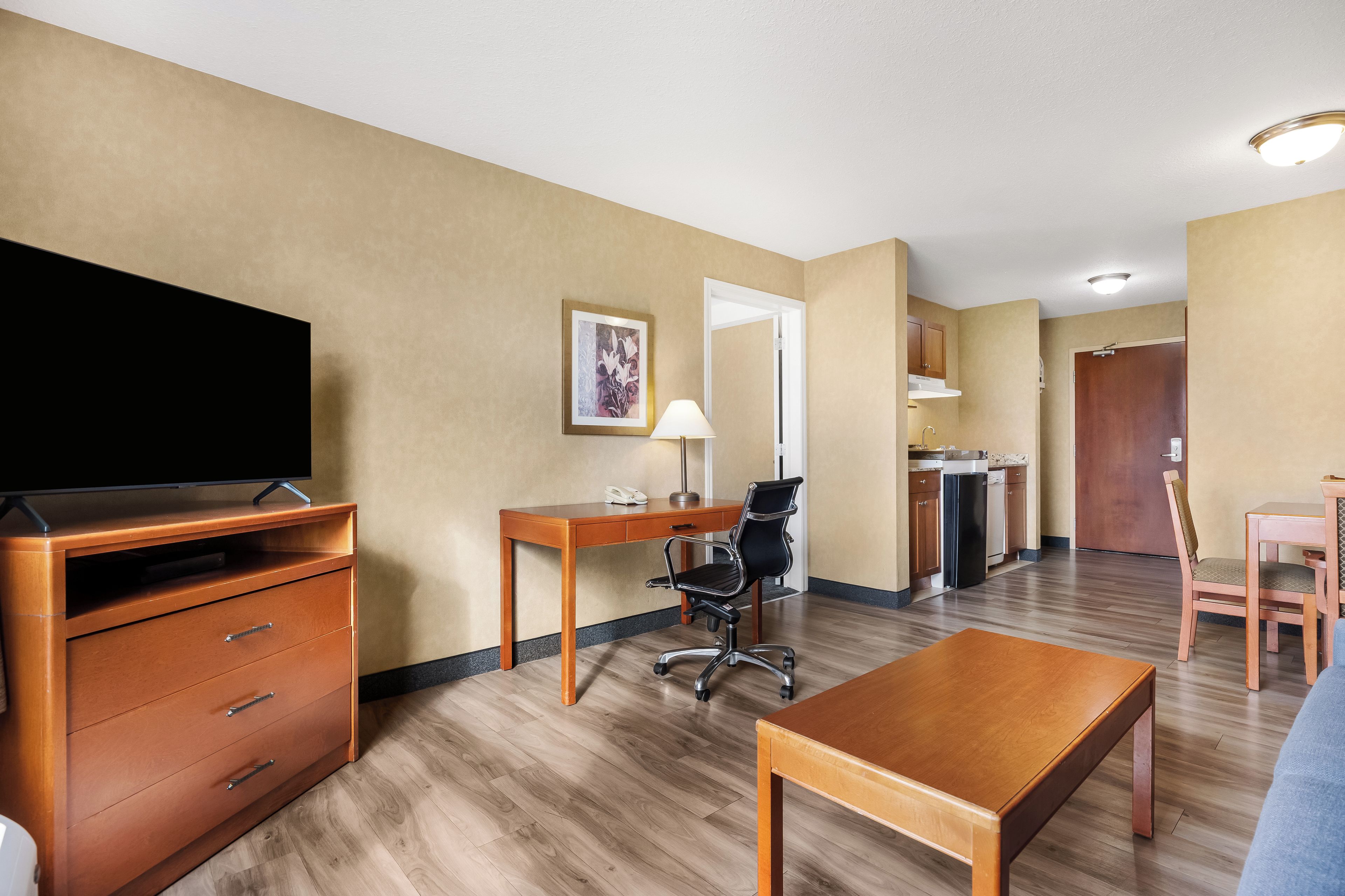 A hotel room with a wooden desk, a chair, a television, a bed, and a kitchenette.